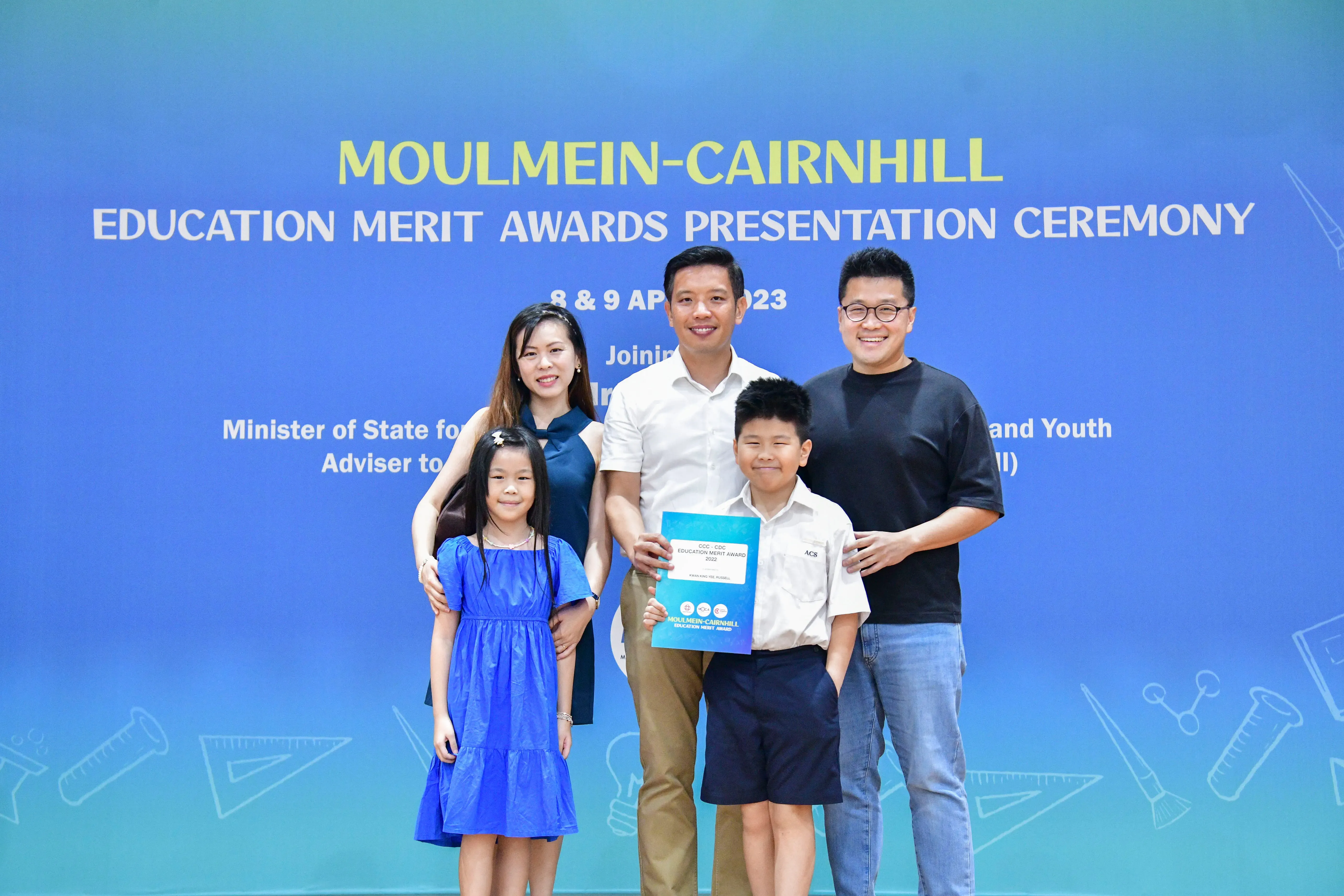 Five people pose before a Moulmein-Cairnhill Education Merit Awards Presentation Ceremony banner, with a boy holding an award.