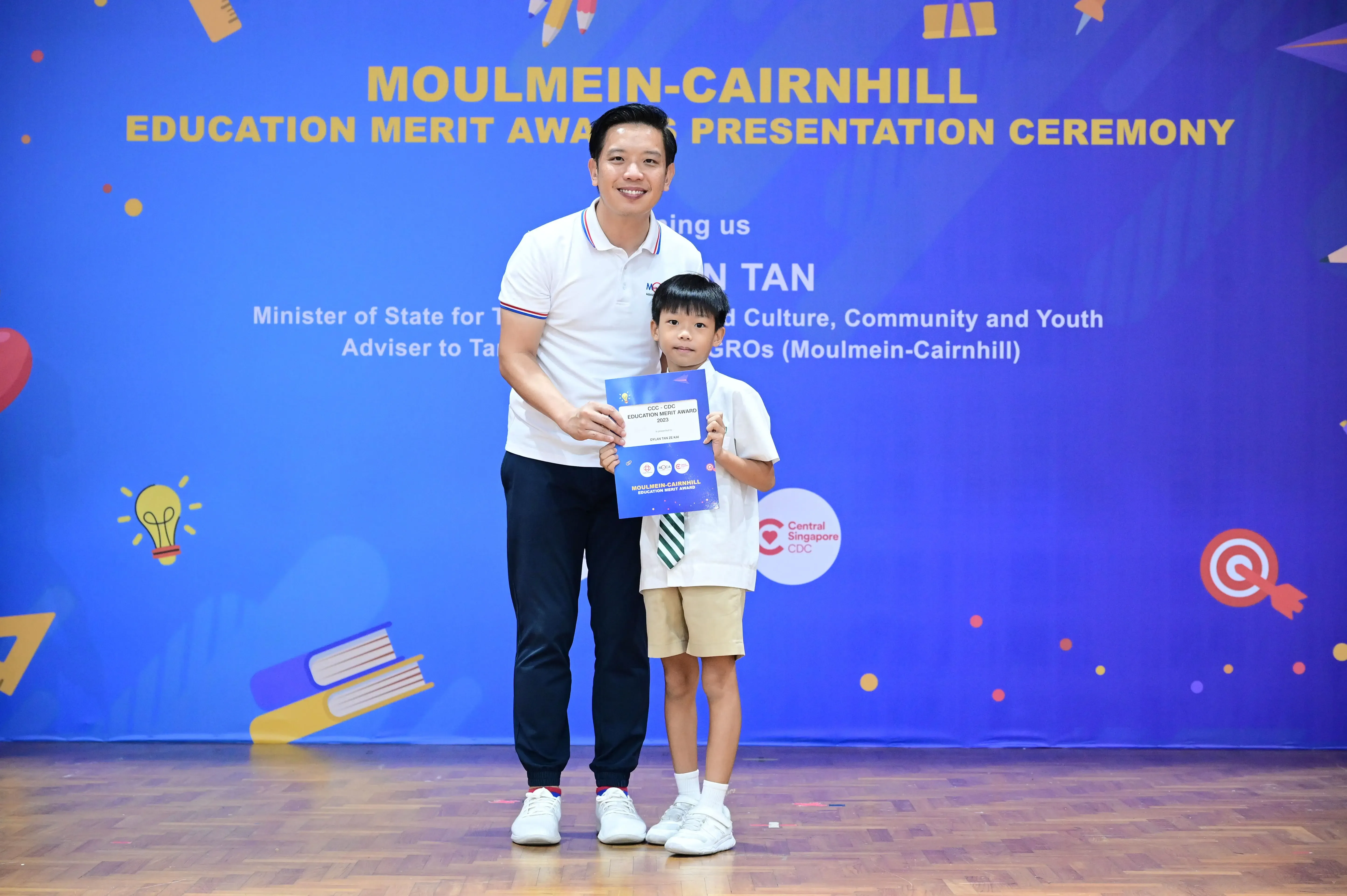 A man and a young boy hold a certificate on stage at the Moulmein-Cairnhill Education Merit Award Presentation Ceremony.