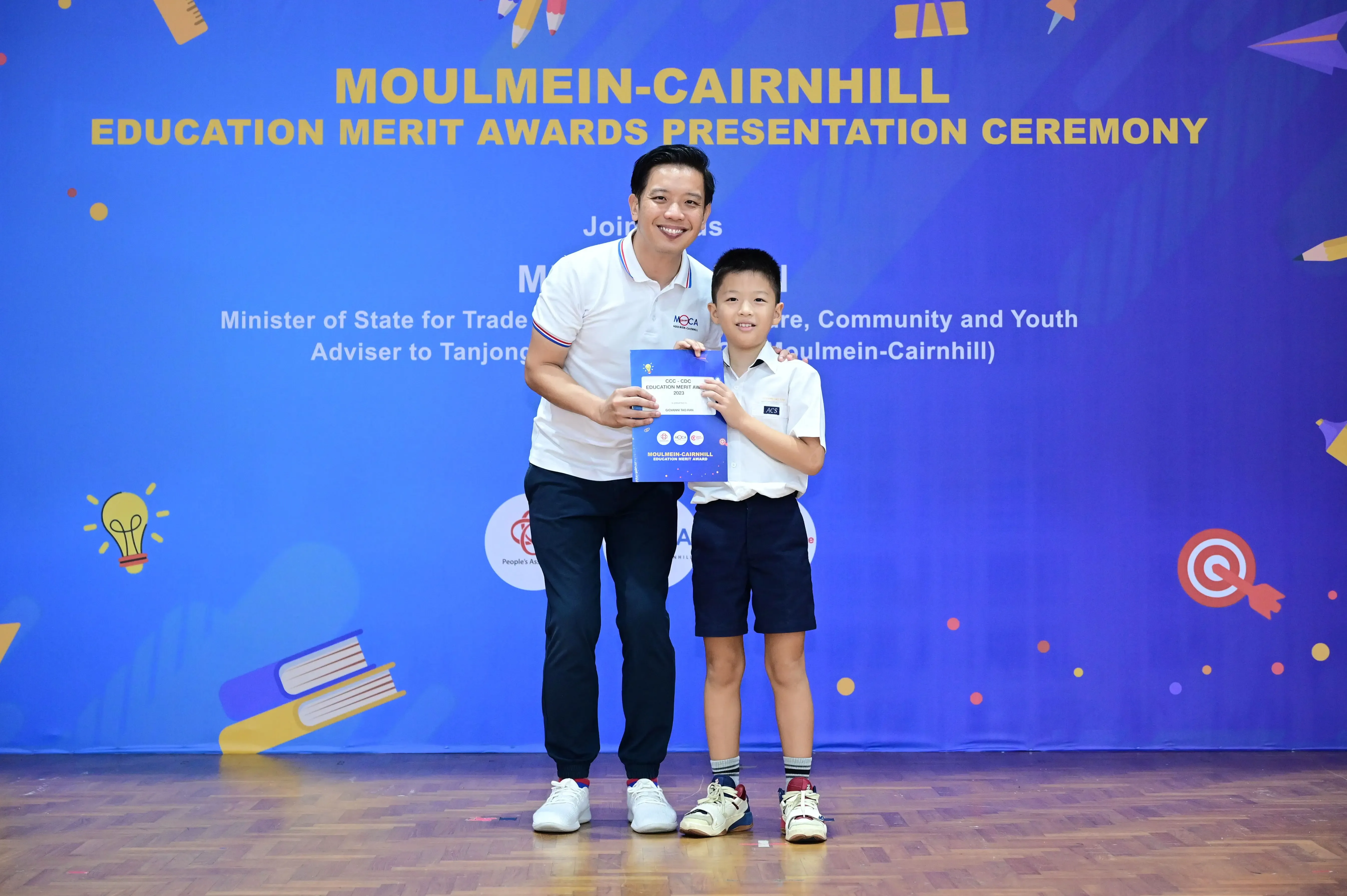 An adult and a young boy hold a merit award certificate at the Moulmein-Cairnhill Education Merit Awards ceremony.