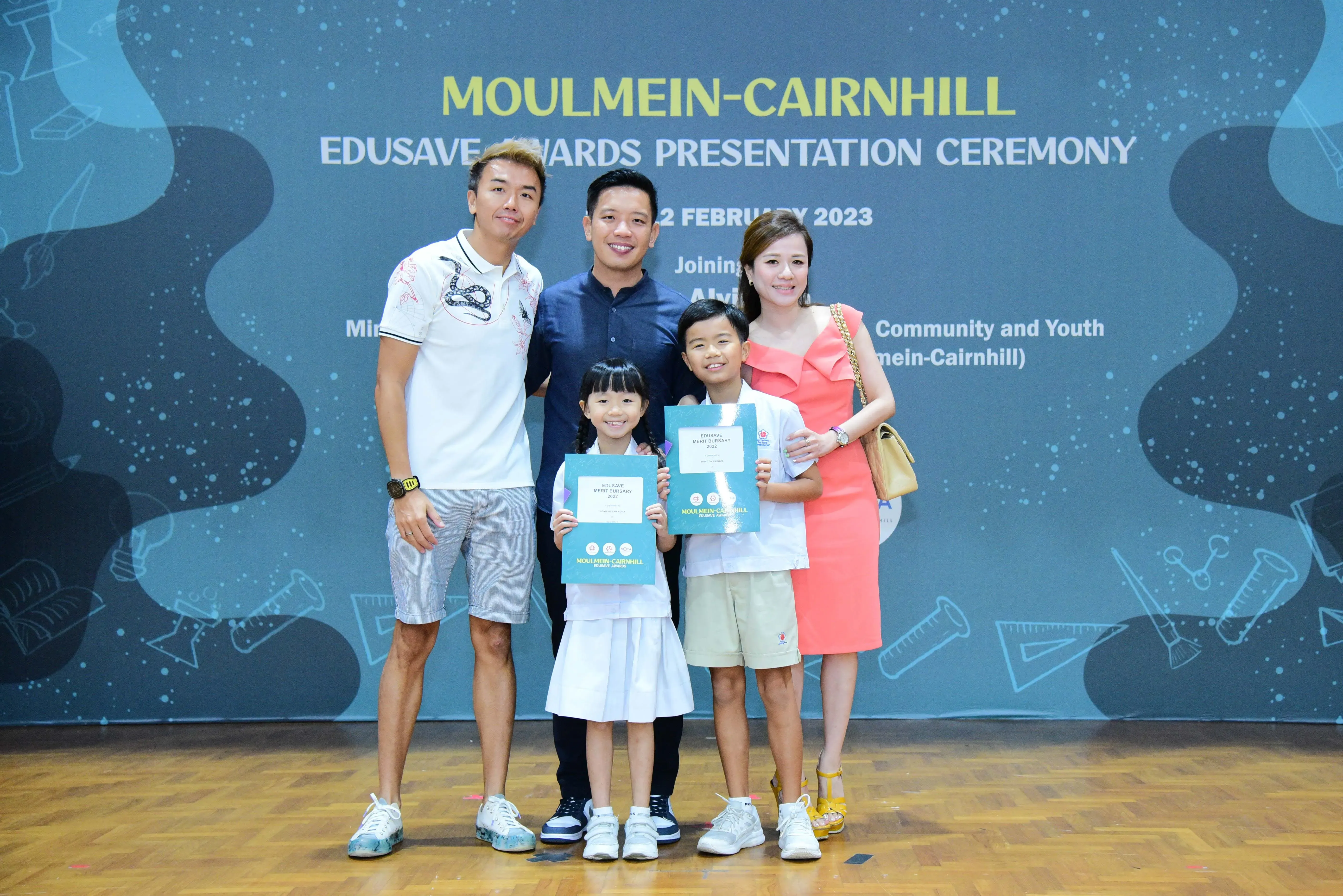 Five people pose at the Moulmein-Cairnhill Edusave Awards Ceremony; two children hold teal award certificates.