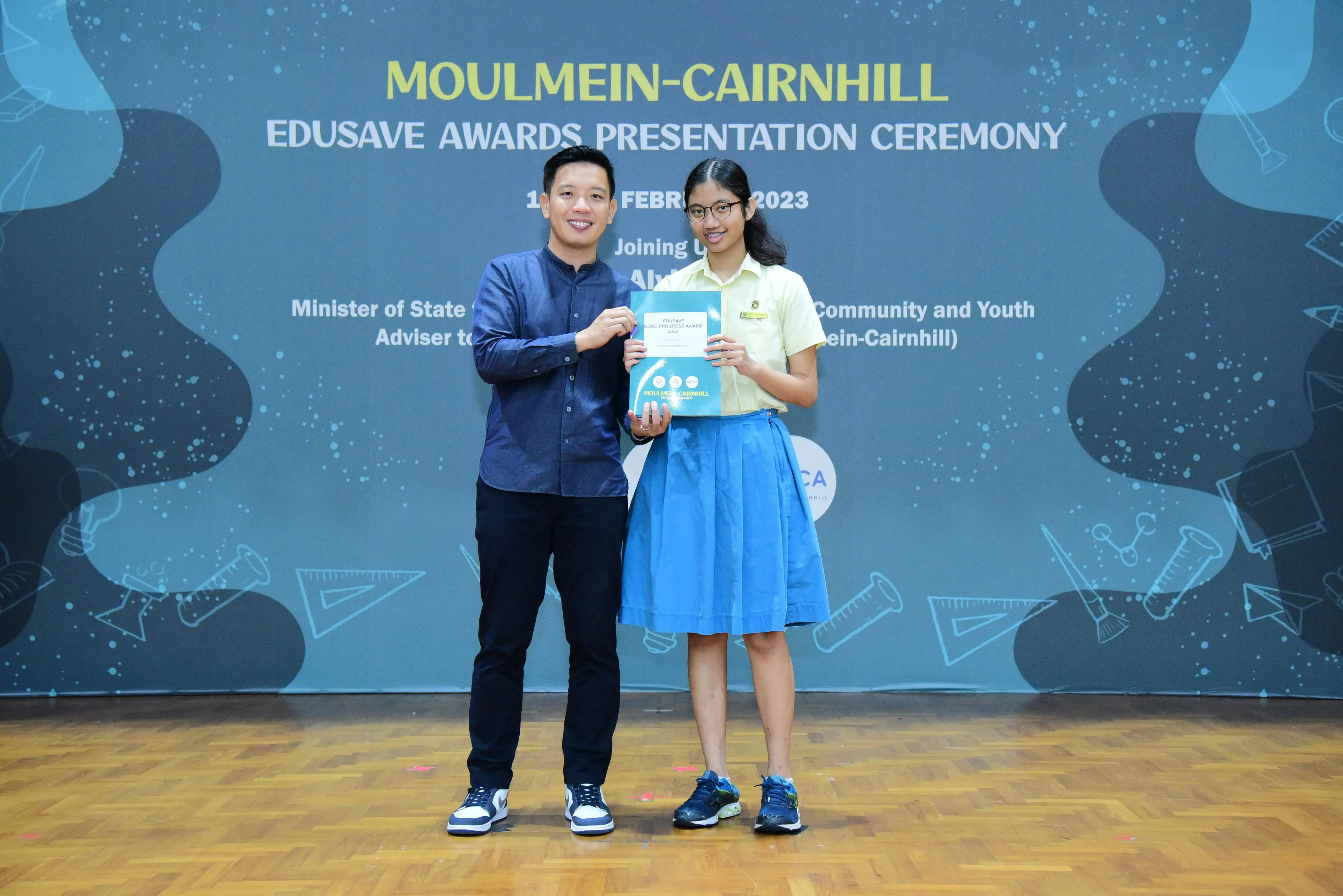 Two people hold a certificate together on stage at the Moulmein-Cairnhill Edusave Awards Presentation Ceremony 2023.