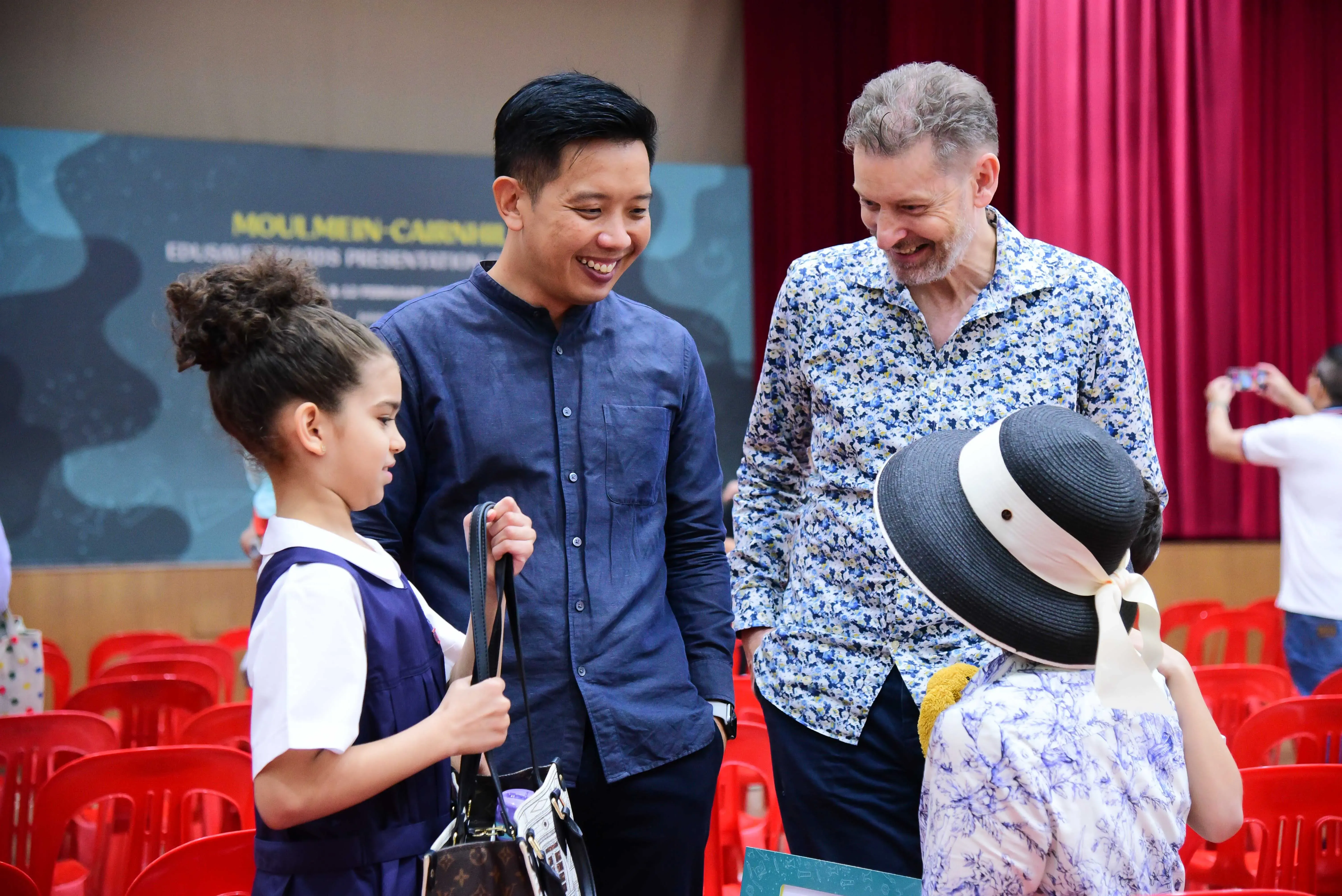 Two smiling men talk with two young children in a hall with red chairs and a presentation banner in the background.
