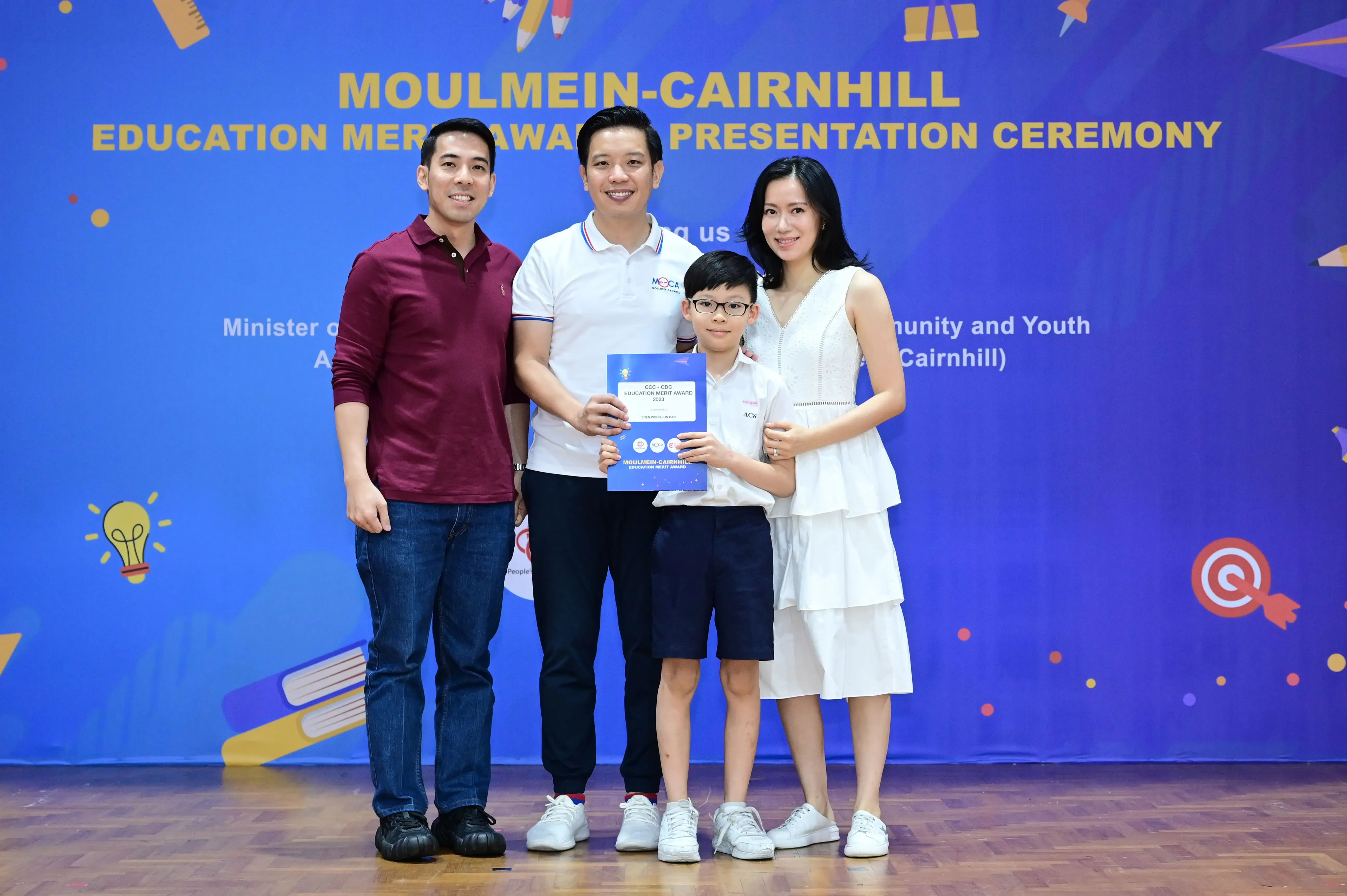 Four people pose before a Moulmein-Cairnhill Education Merit Award Presentation Ceremony banner, a boy holding a certificate.