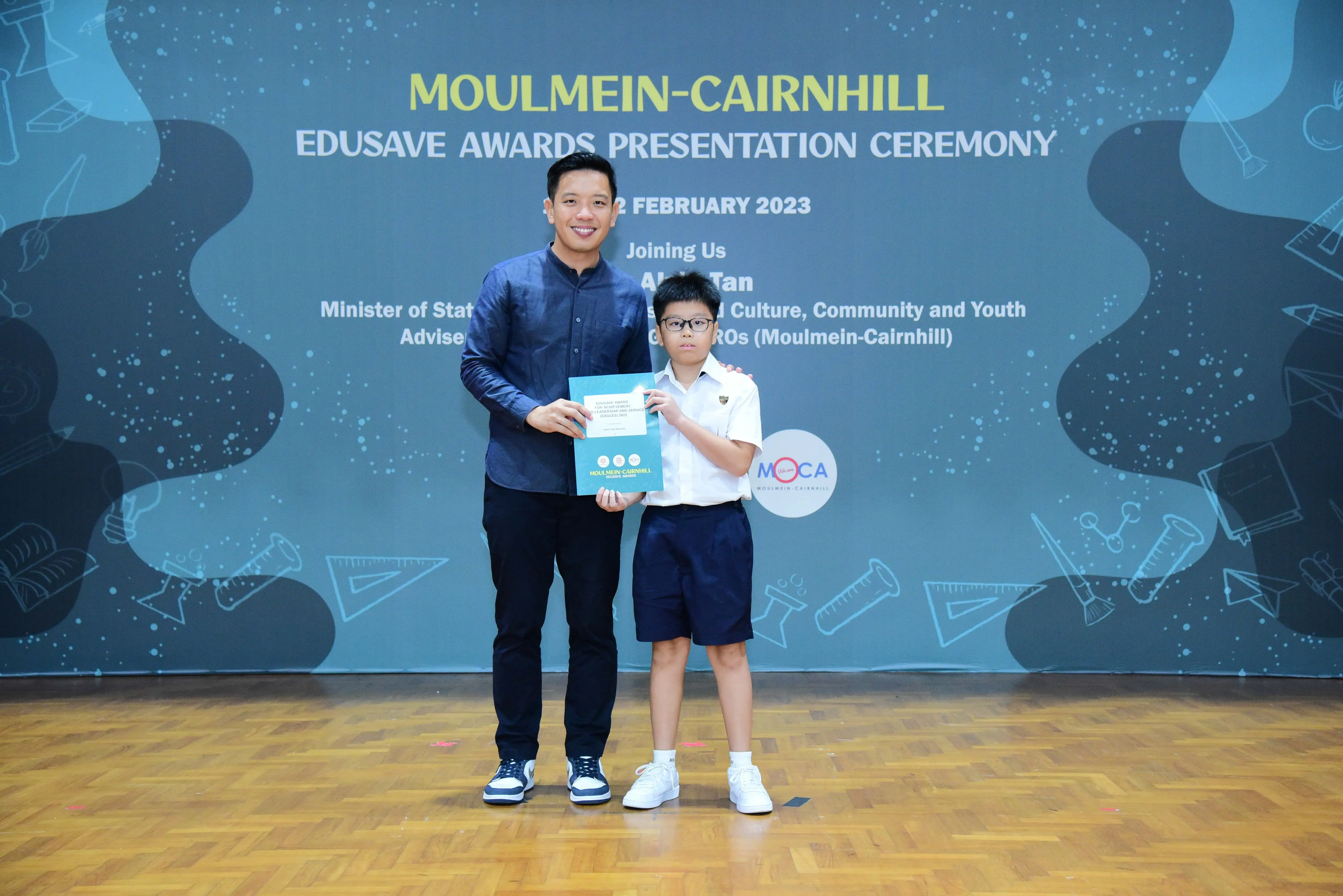 An adult and a young student hold a certificate at the Moulmein-Cairnhill Edusave Awards Ceremony, February 2023.