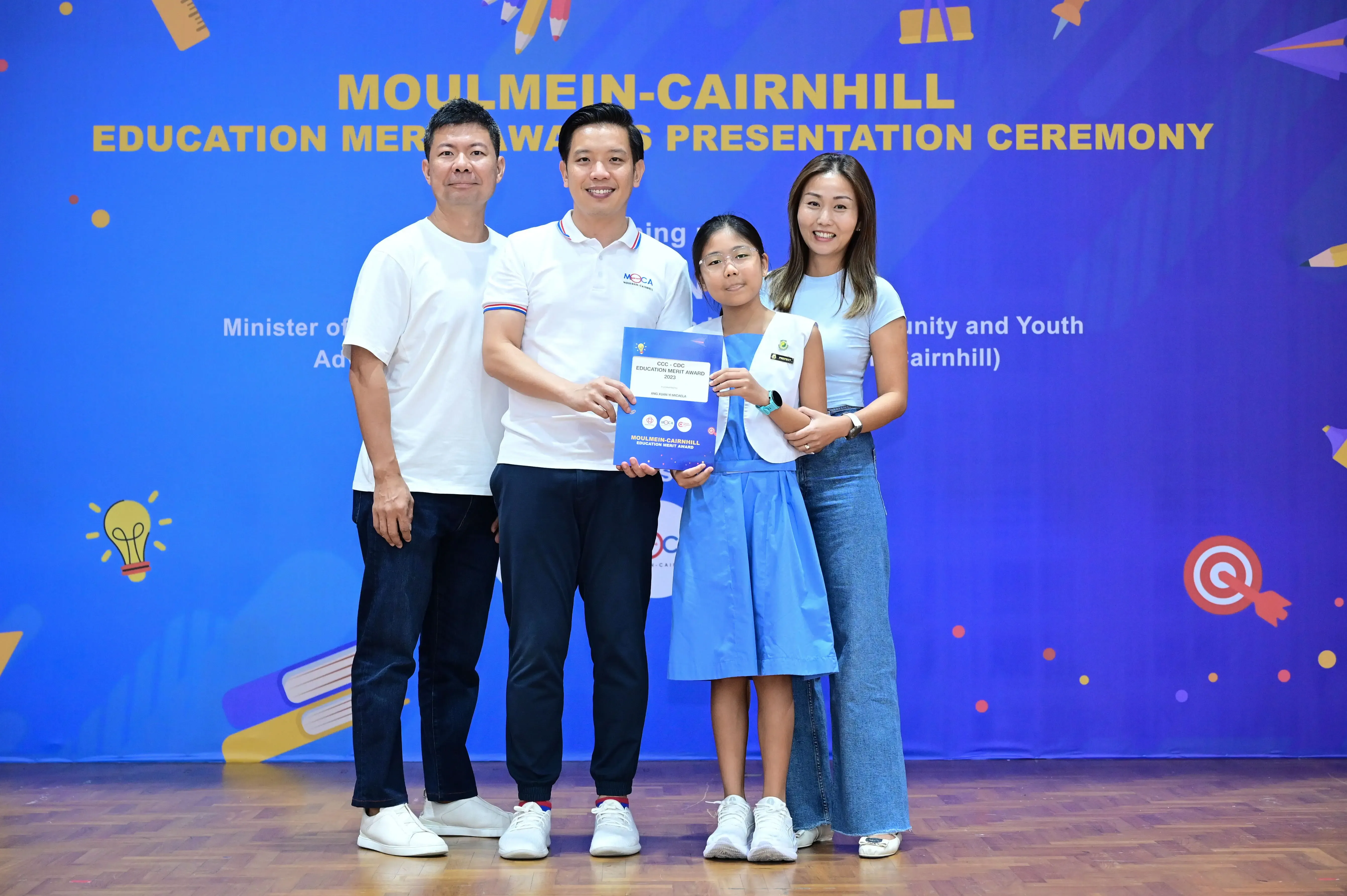 Four people pose at the Moulmein-Cairnhill Education Merit Awards Presentation Ceremony, with a girl holding an award certificate.