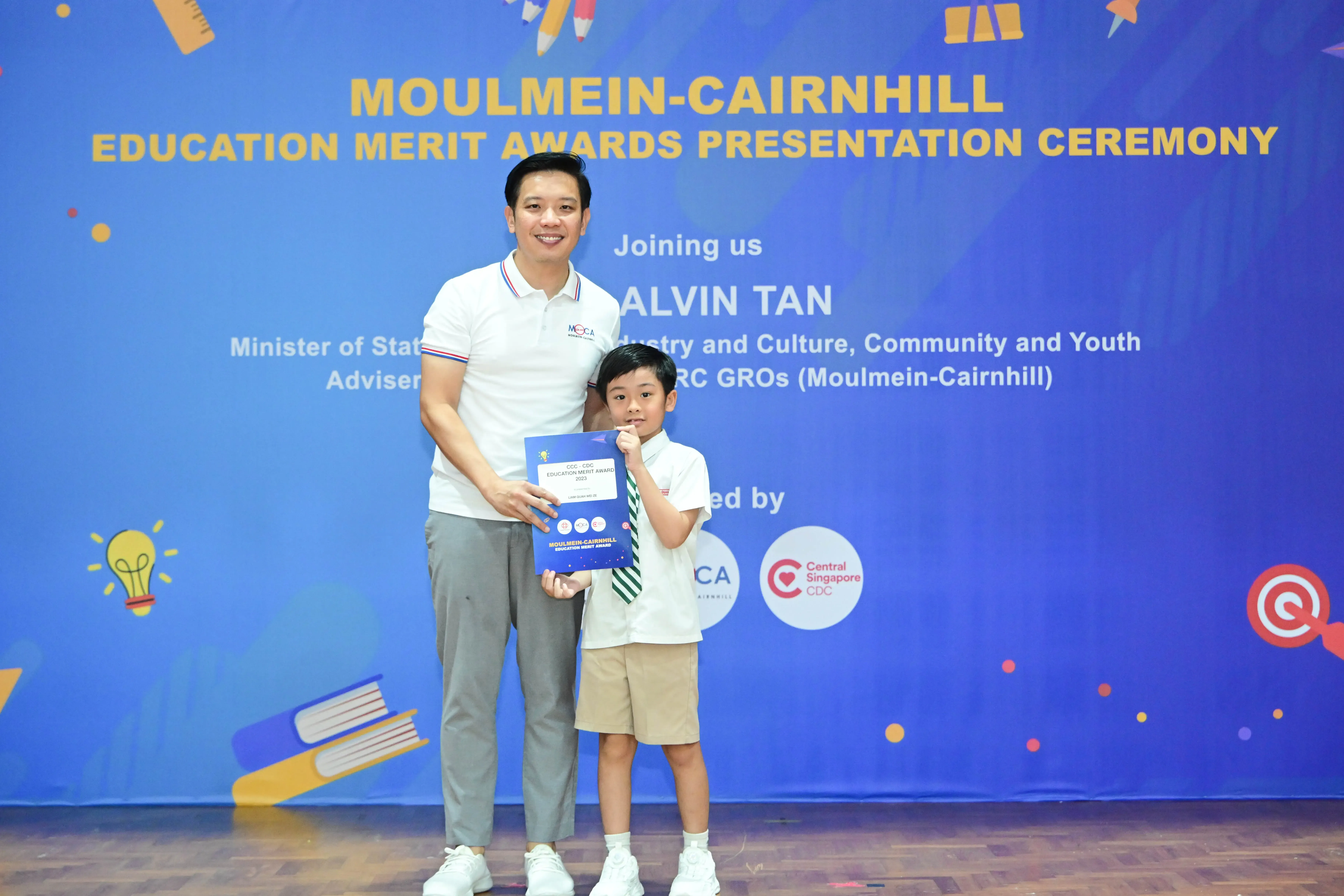 A man and a young boy hold a blue award certificate in front of the Moulmein-Cairnhill Education Merit Awards banner.