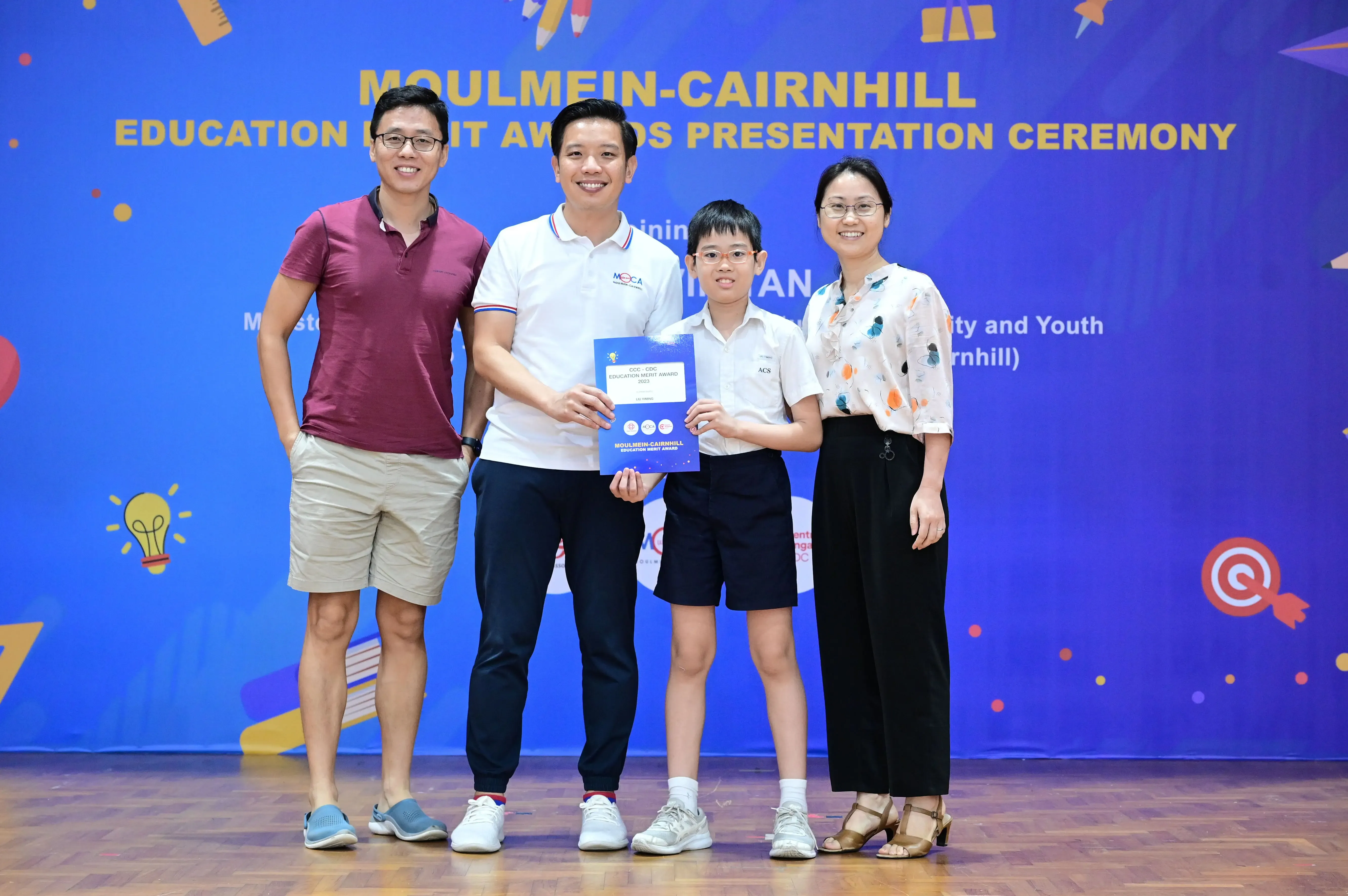 Four people pose at the Moulmein-Cairnhill Education Merit Awards Presentation Ceremony, with a boy holding an award certificate.