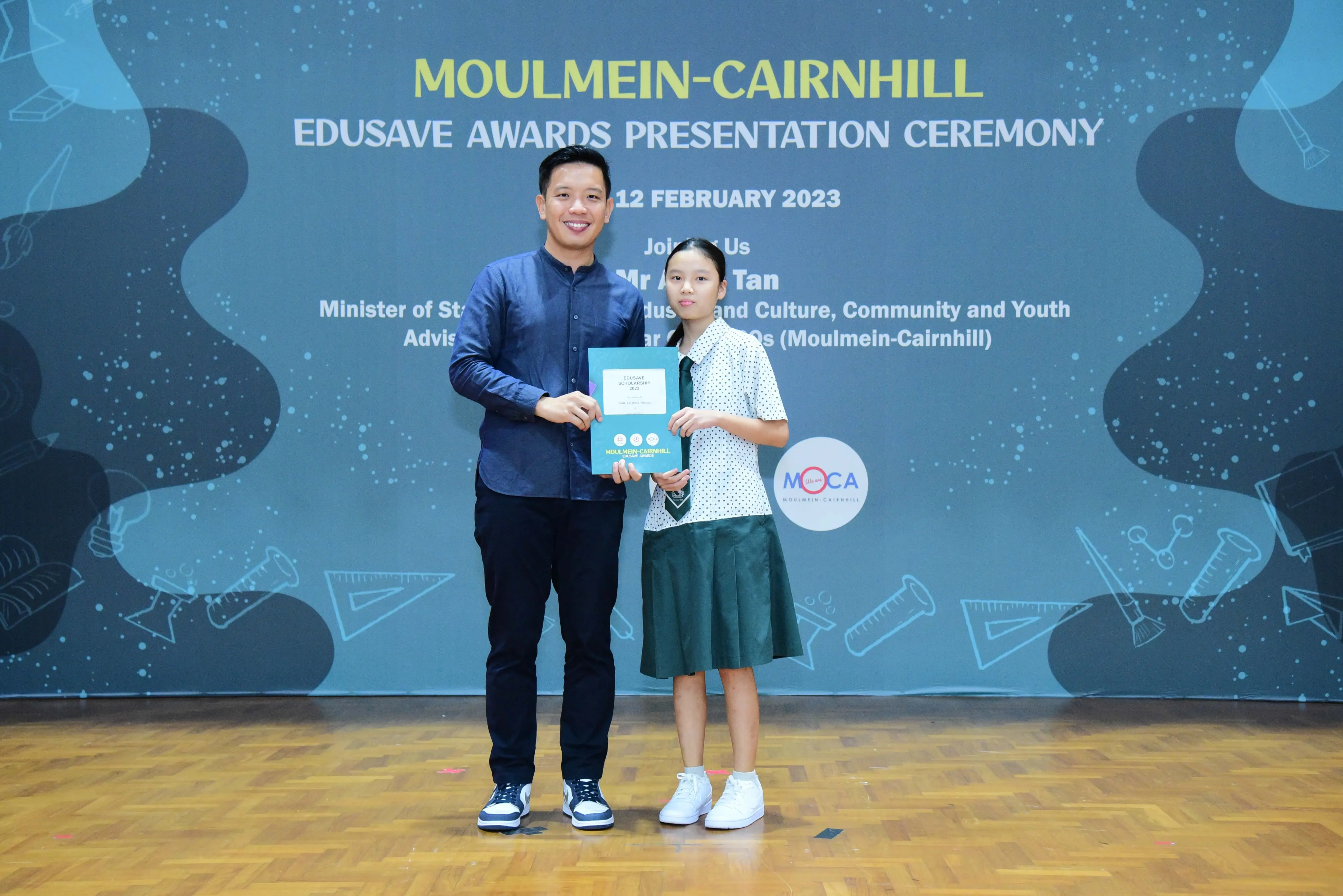 A man and a young girl in school uniform hold a certificate at the Moulmein-Cairnhill Edusave Awards Ceremony, February 2023.