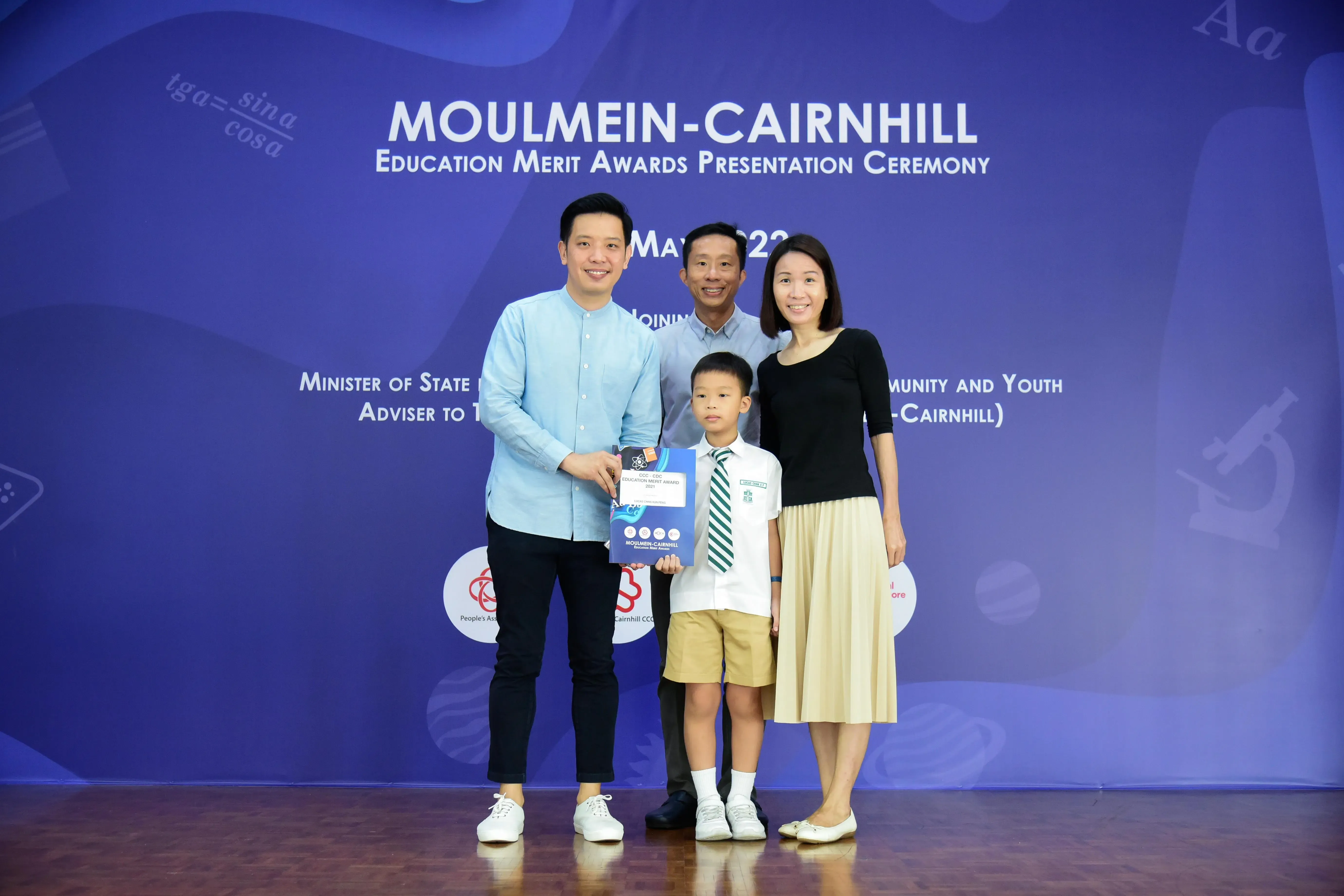 Four people pose before a Moulmein-Cairnhill Education Merit Awards banner, a young boy holding a blue award folder.