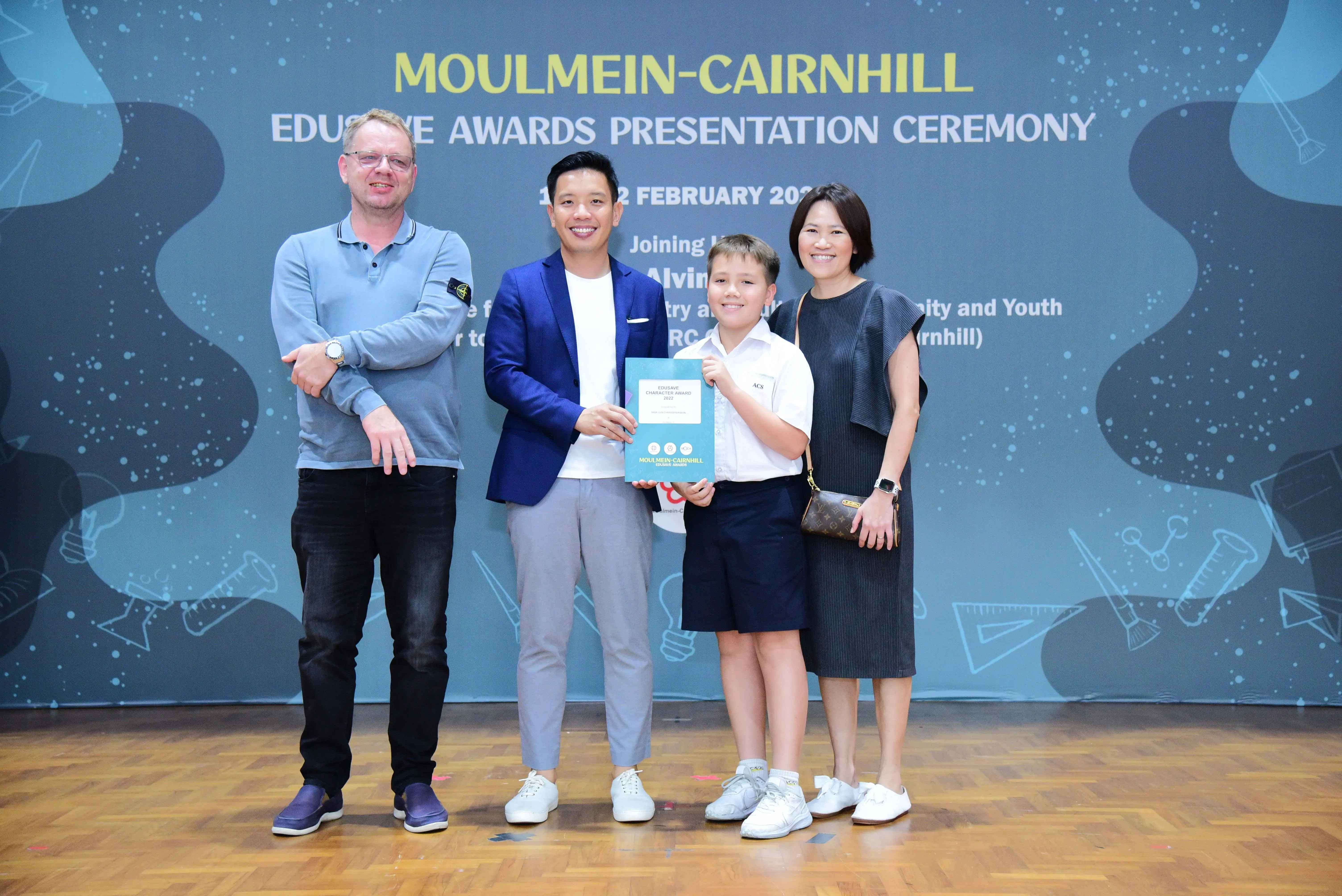 Four people pose at the Moulmein-Cairnhill Edusave Awards Presentation Ceremony, with a boy holding a certificate.