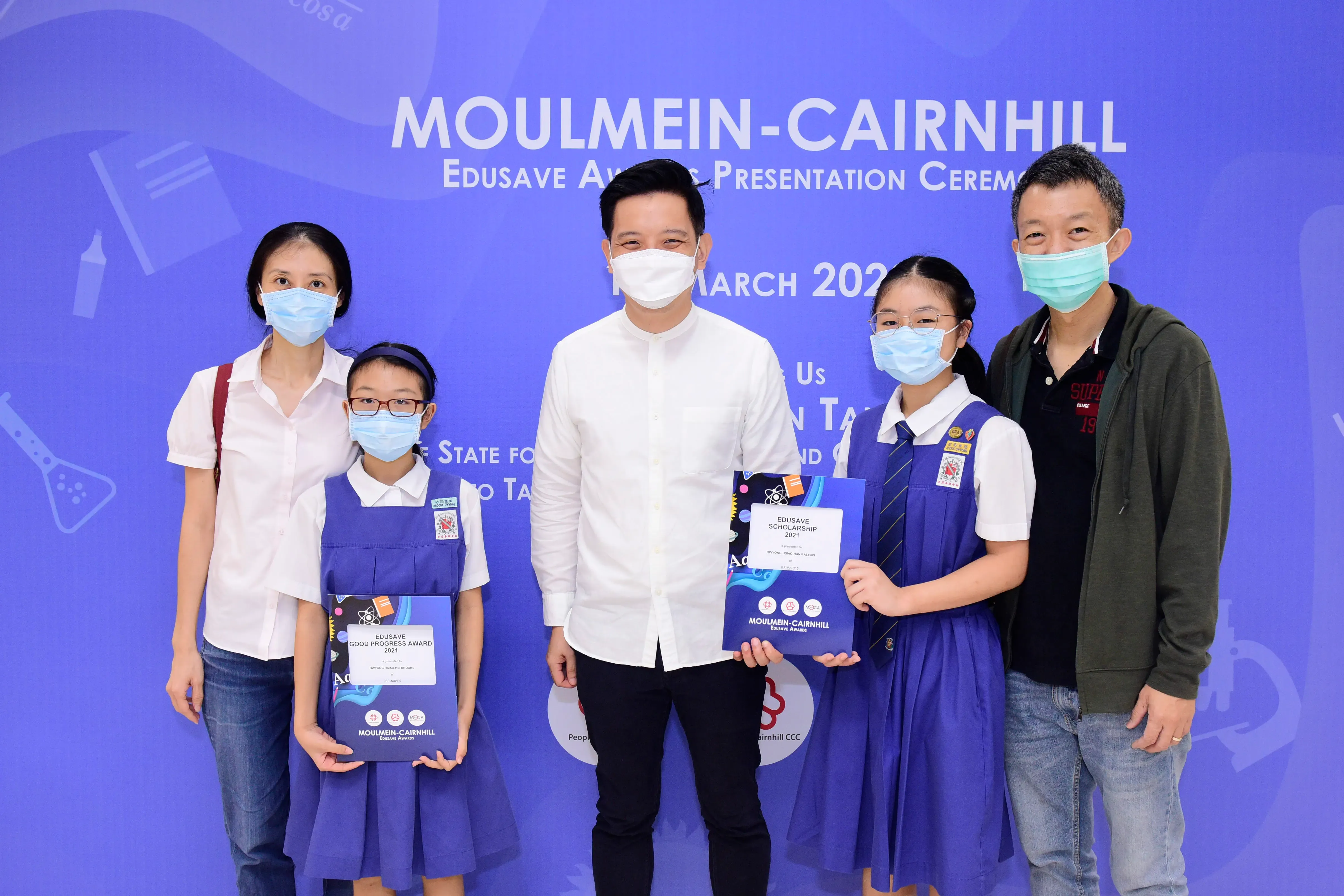 Five masked people pose at the Moulmein-Cairnhill Edusave Awards Ceremony, two girls holding award certificates.