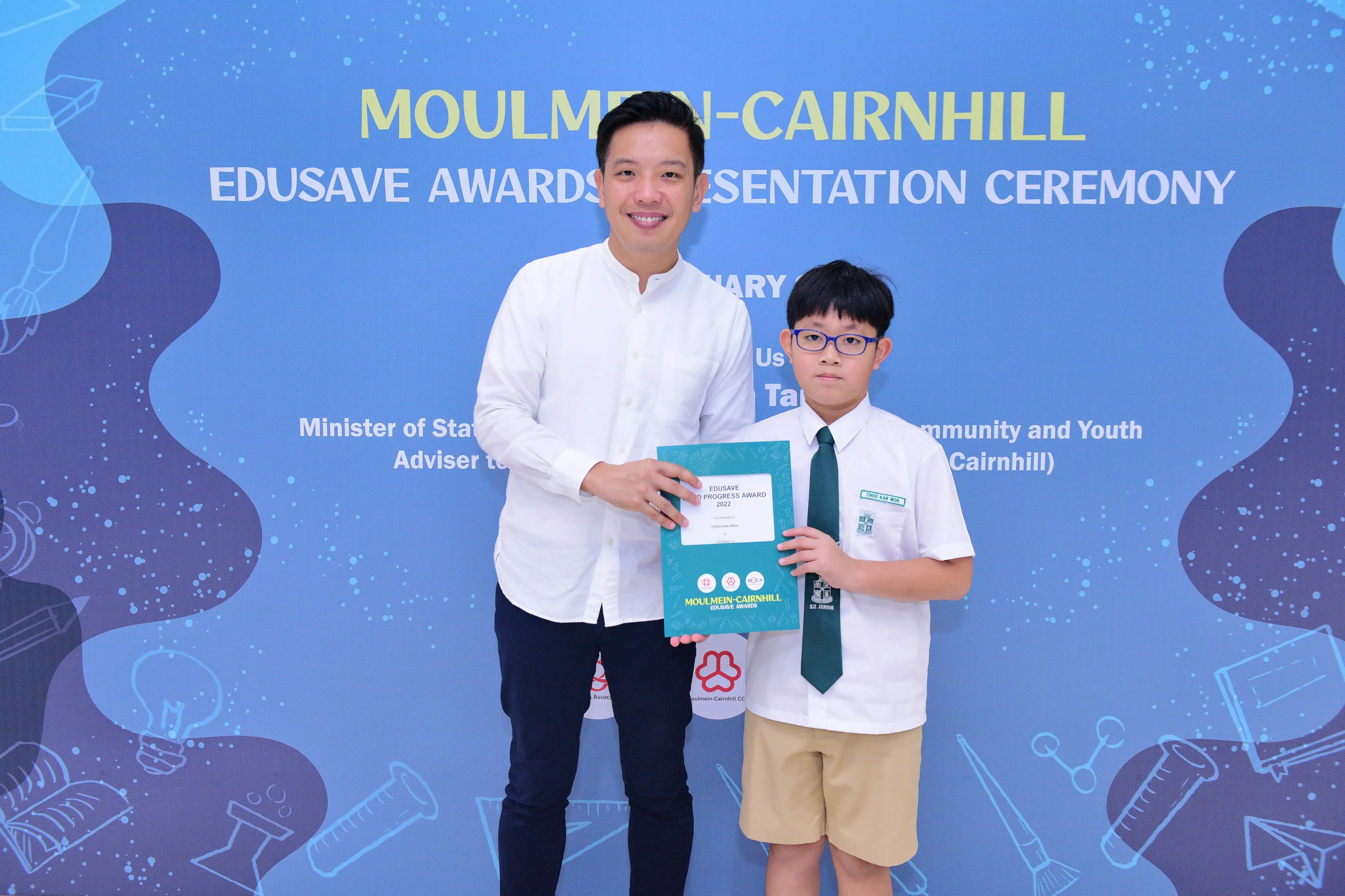 An adult and a young student hold a teal award folder at the Moulmein-Cairnhill Edusave Awards Presentation Ceremony.