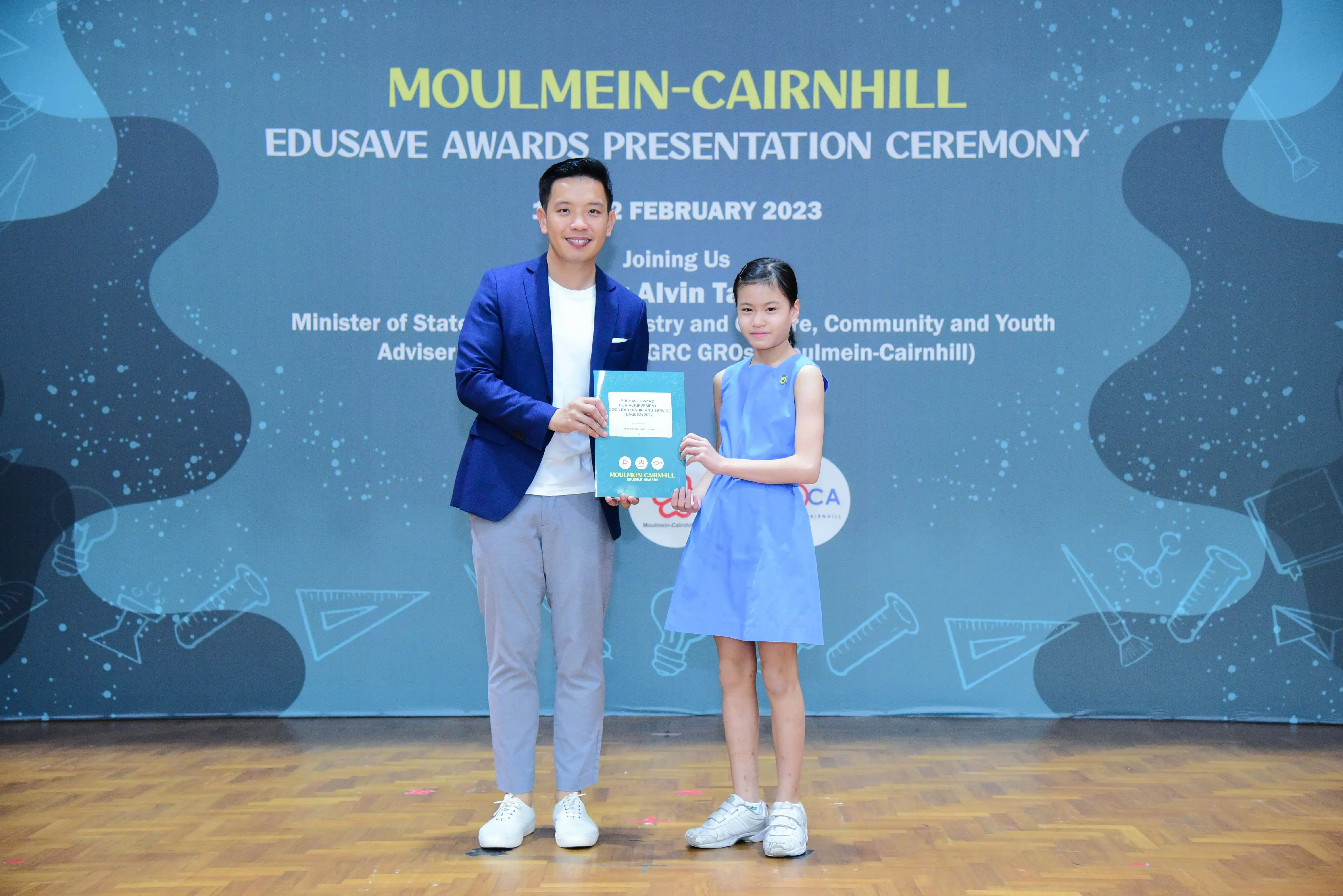 A man in a blue blazer and a young girl in a blue dress hold a certificate at the Moulmein-Cairnhill Edusave Awards 2023.