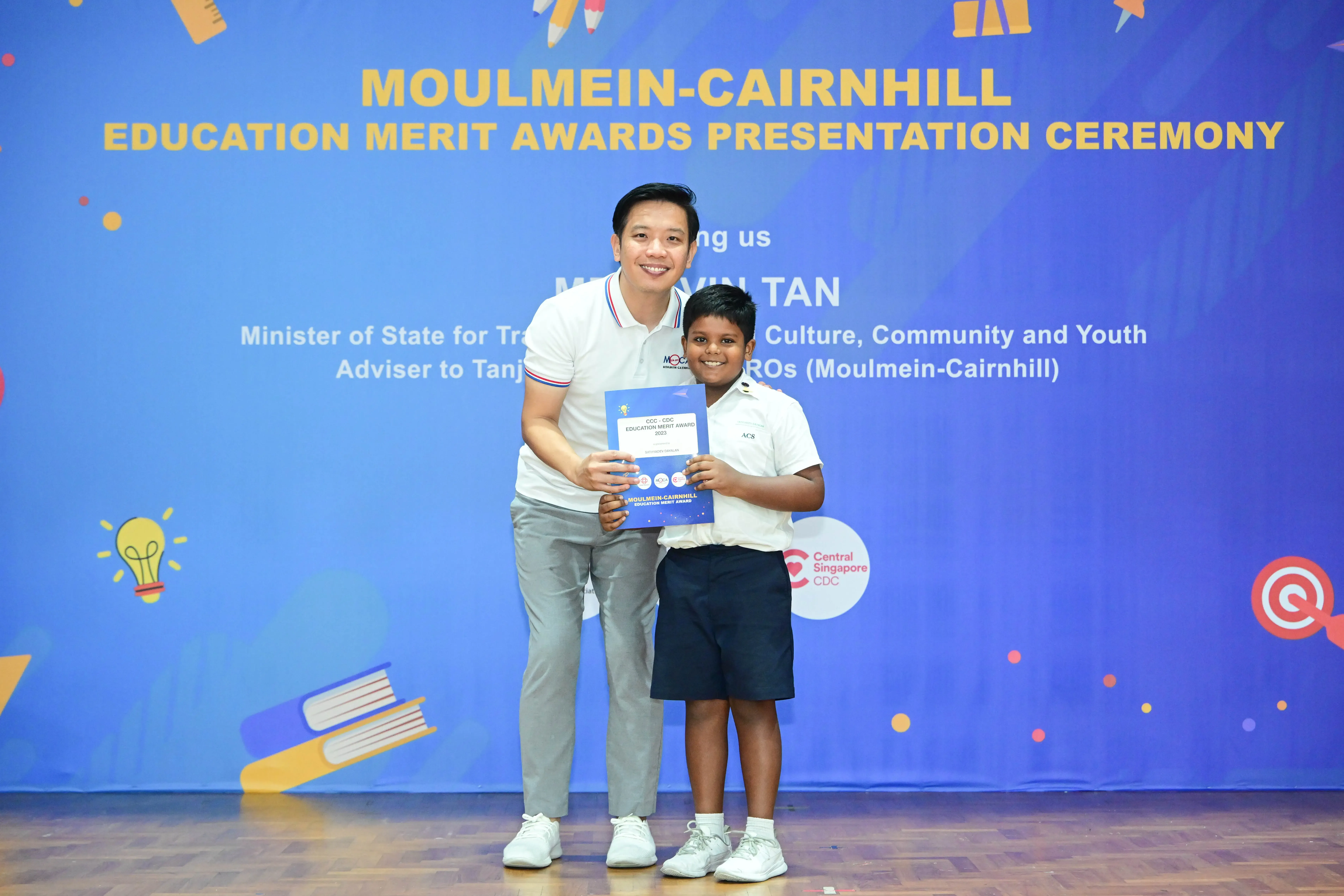 A man and a young boy hold a certificate together in front of a Moulmein-Cairnhill Education Merit Awards banner.