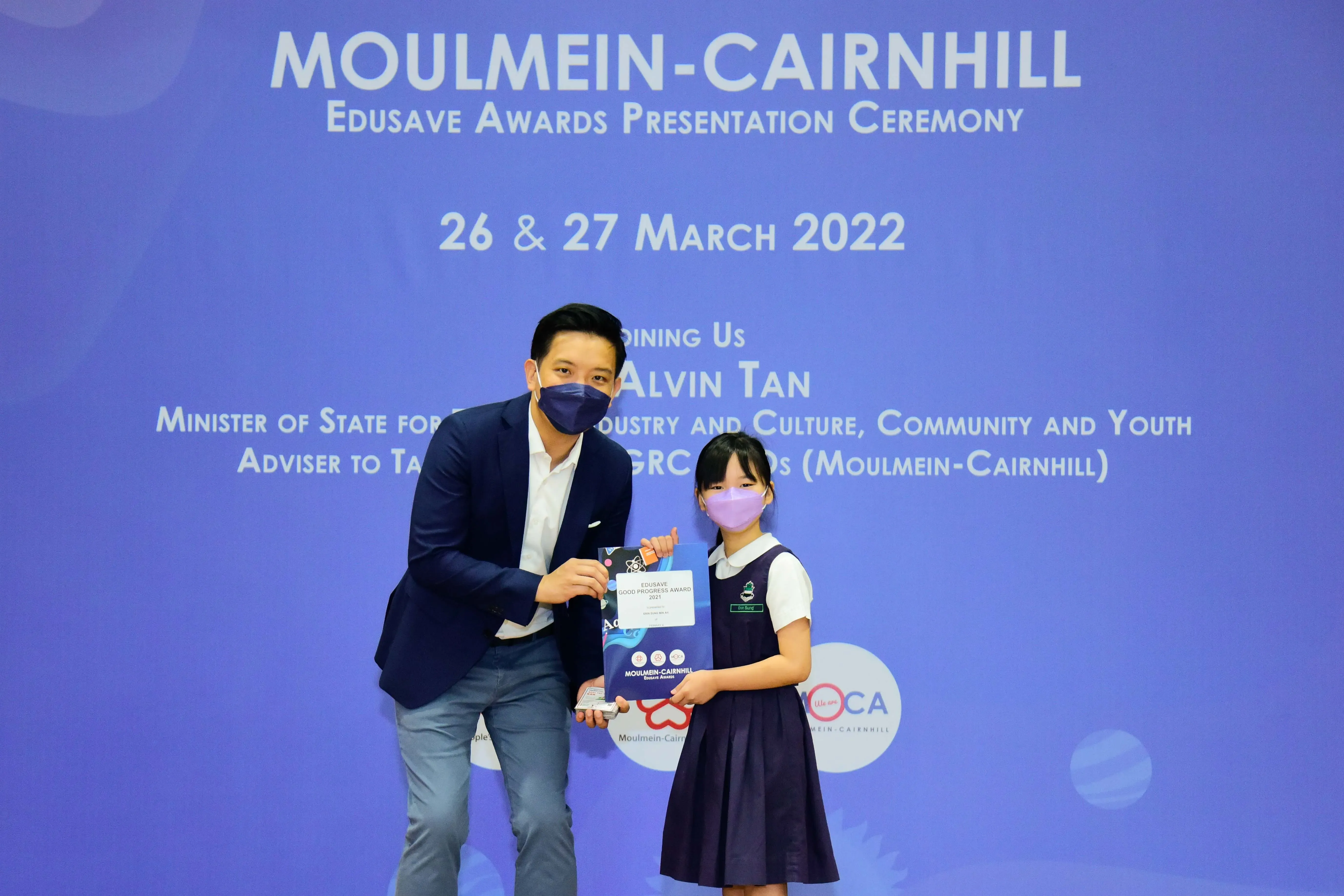 A masked man in a blazer presents an award to a young masked girl at the Moulmein-Cairnhill Edusave Awards Ceremony 2022.