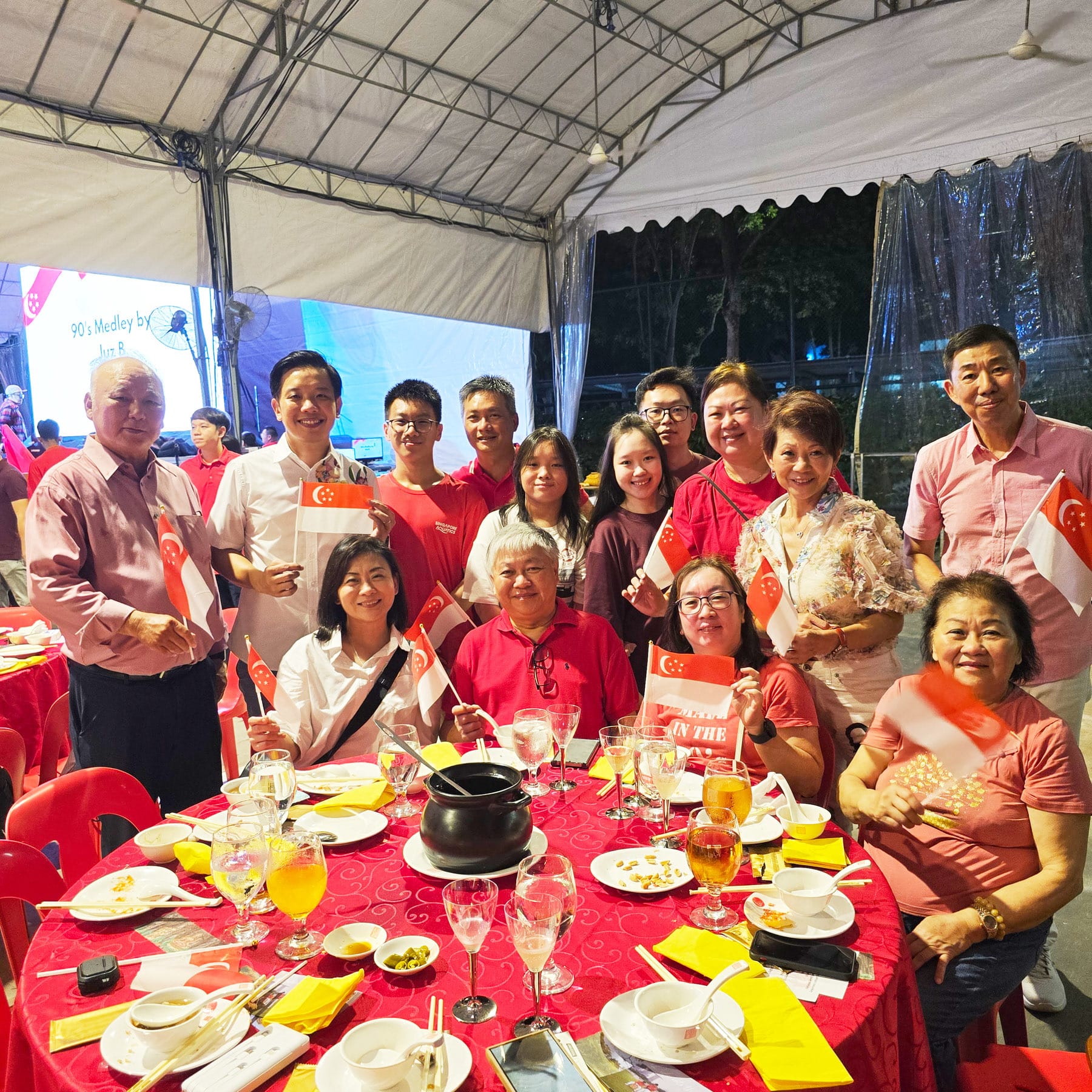 Group of people in red attire holding Singapore flags at a festive dinner under a tent with a red tablecloth.