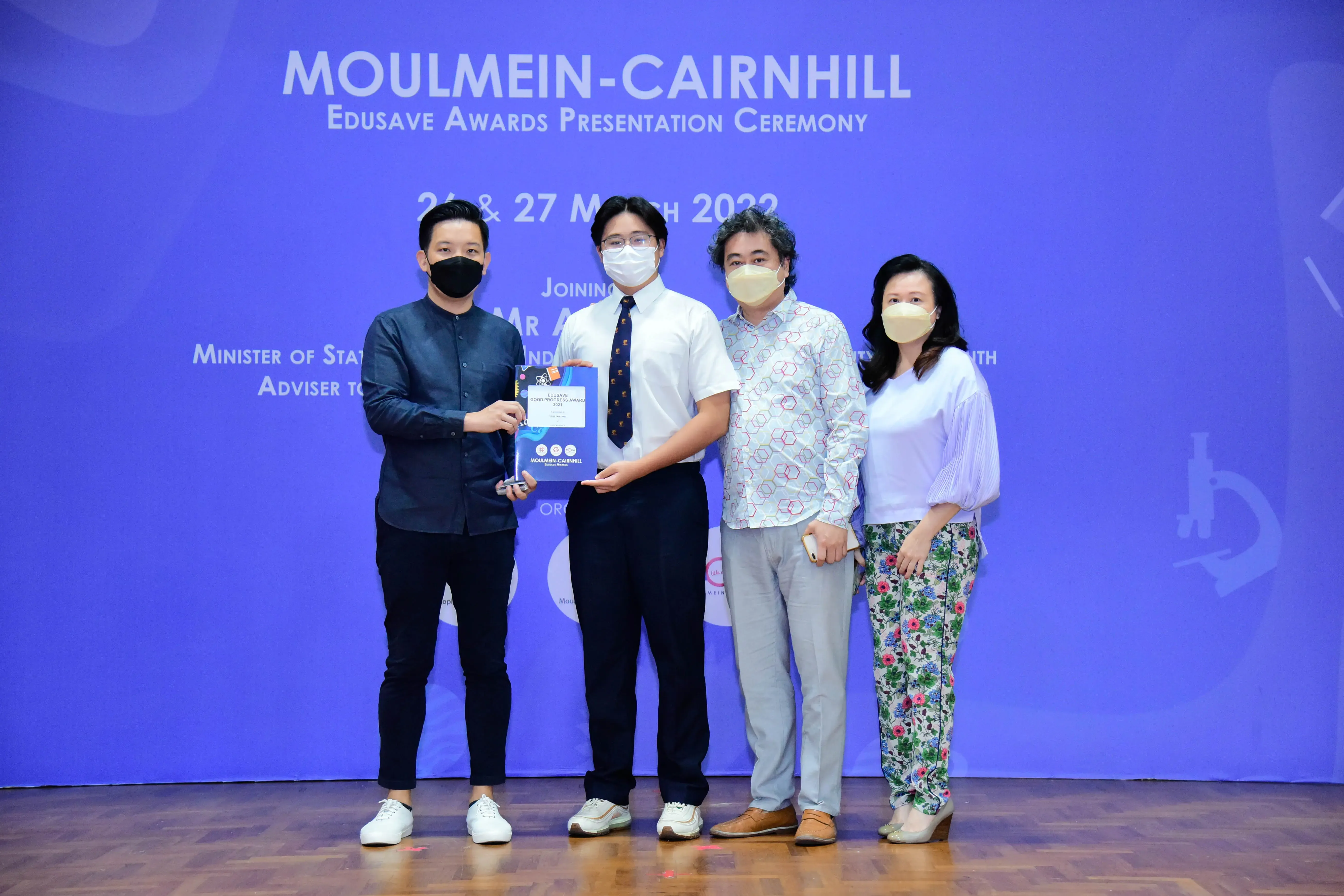Four masked people pose at the Moulmein-Cairnhill Edusave Awards Presentation Ceremony, with two holding a certificate.