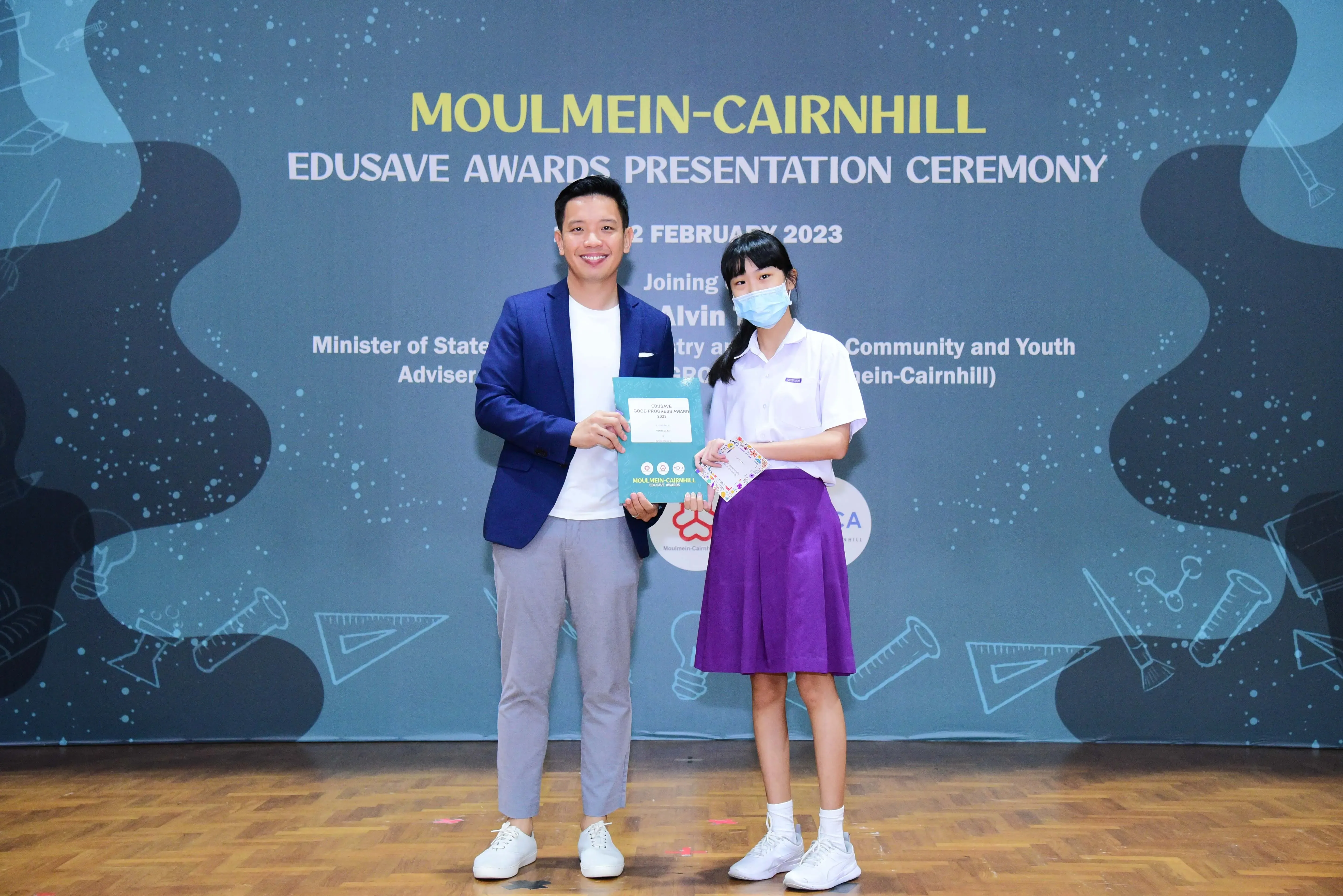 A man in a blue blazer and a masked schoolgirl hold awards at the Moulmein-Cairnhill Edusave Awards Ceremony 2023.