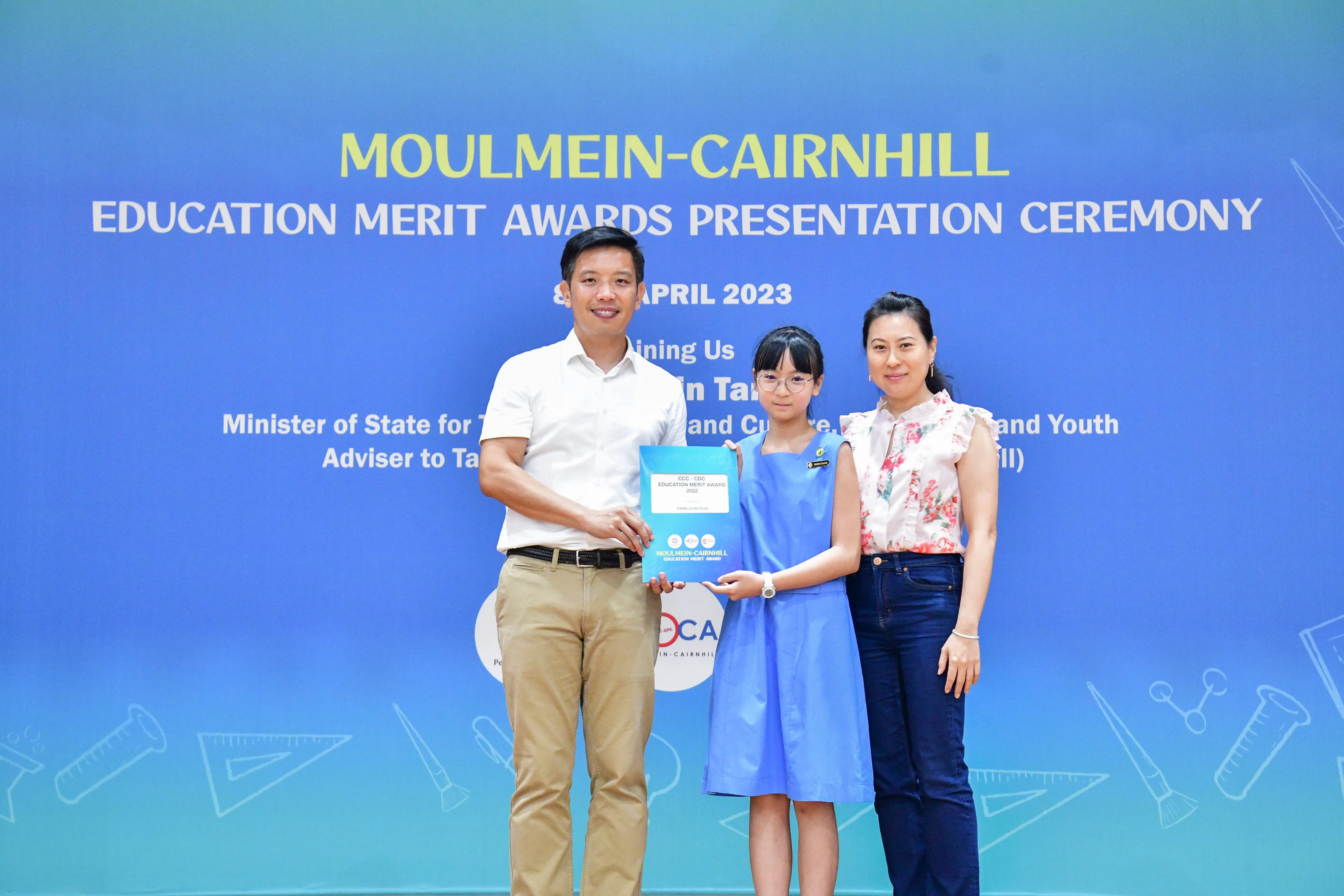 A girl holds an award certificate flanked by two adults at the Moulmein-Cairnhill Education Merit Awards Ceremony, April 2023.