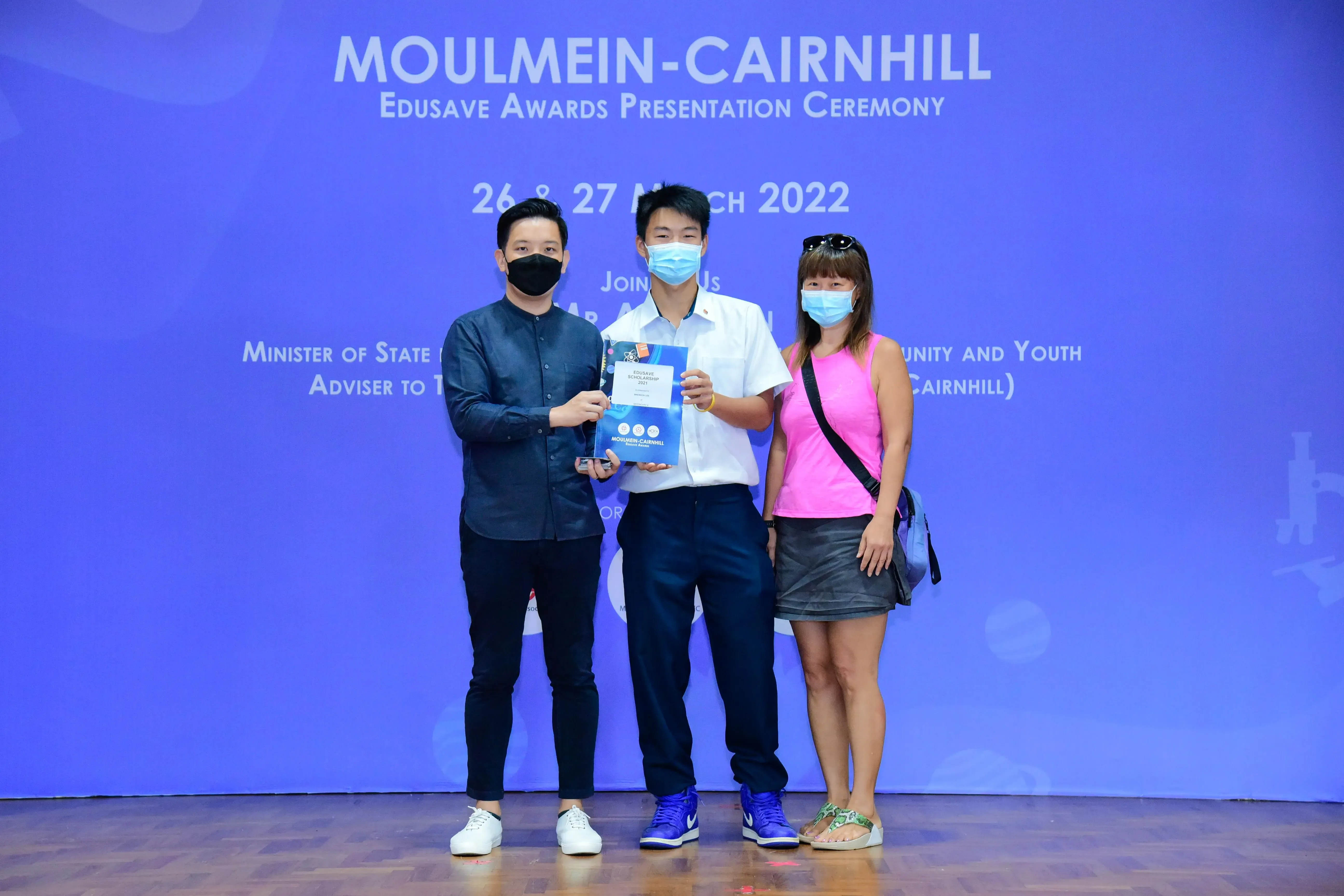 Three masked people pose at the Moulmein-Cairnhill Edusave Awards Presentation Ceremony, with a student holding an award.