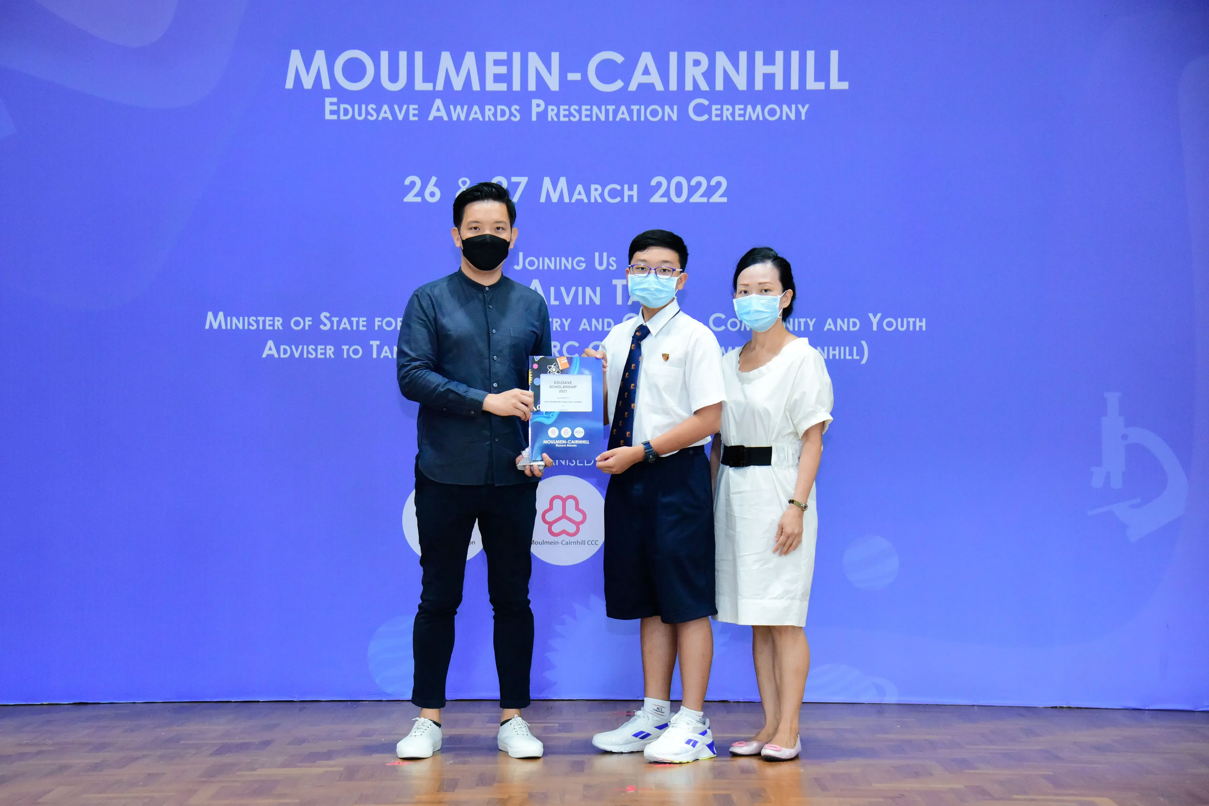 Three masked people stand on a stage holding an award at the Moulmein-Cairnhill Edusave Awards Ceremony, March 2022.