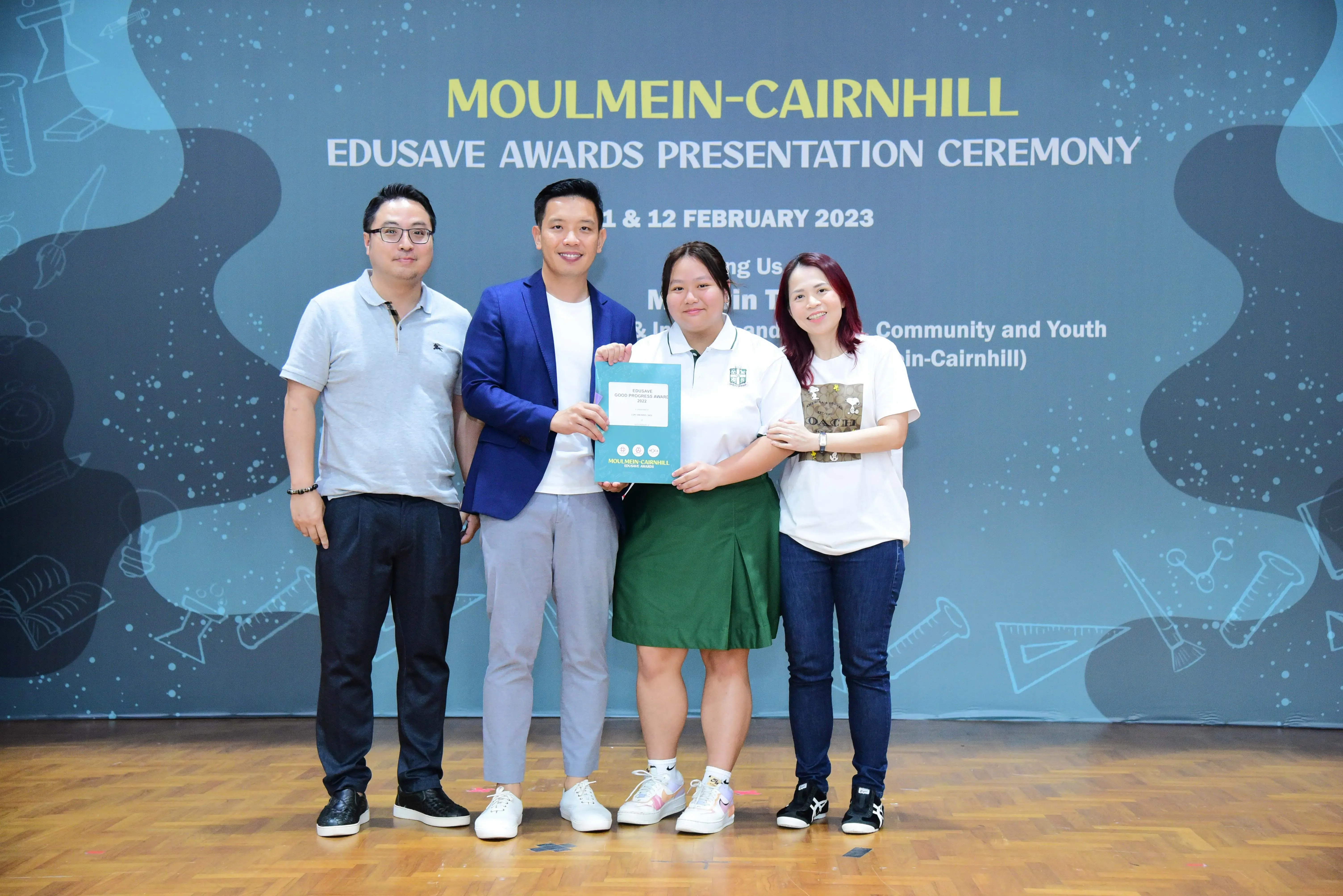 Four people pose on a stage at the Moulmein-Cairnhill Edusave Awards Presentation Ceremony, holding a certificate.