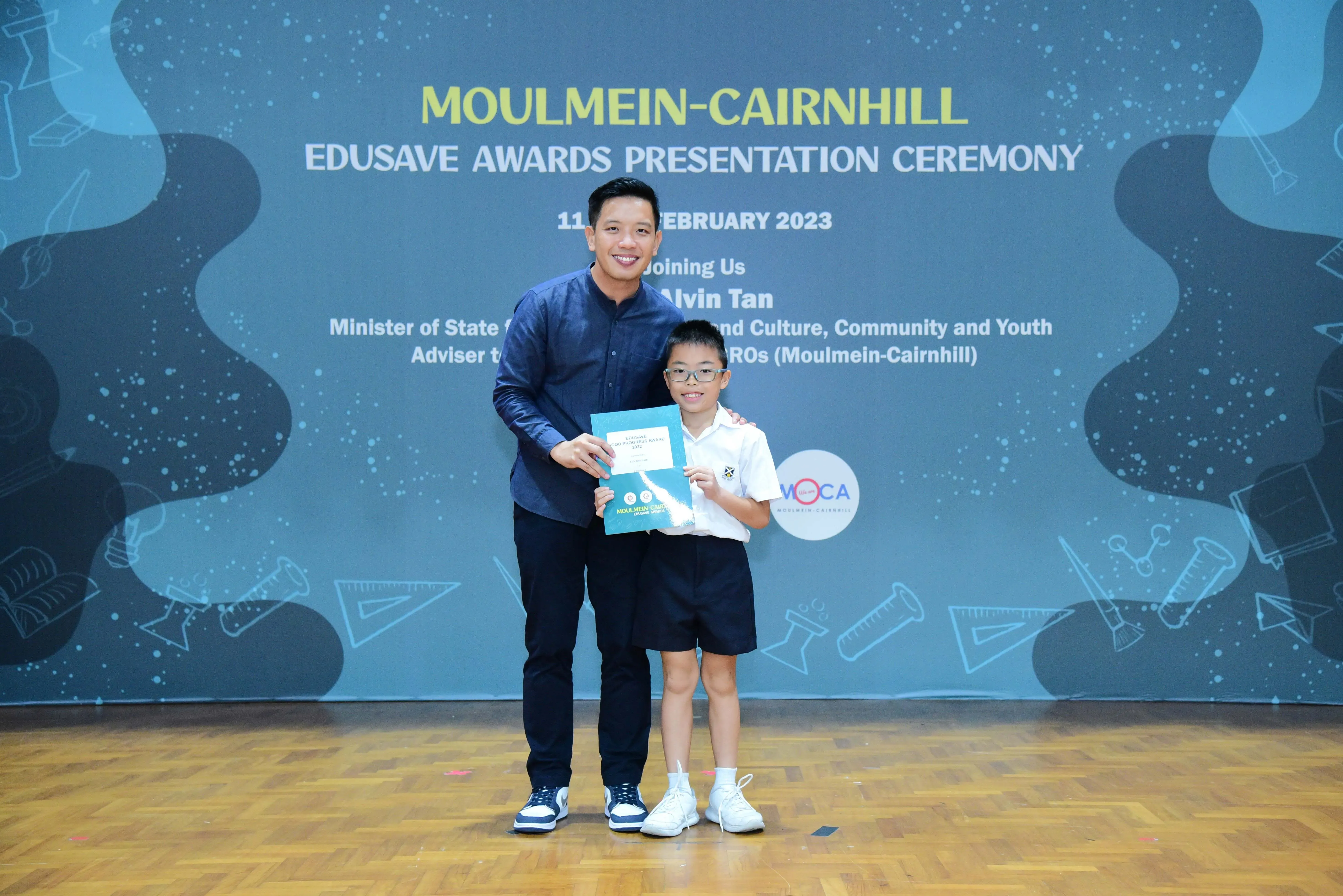 An adult and a young boy in school uniform hold a certificate at the Moulmein-Cairnhill Edusave Awards Ceremony 2023.