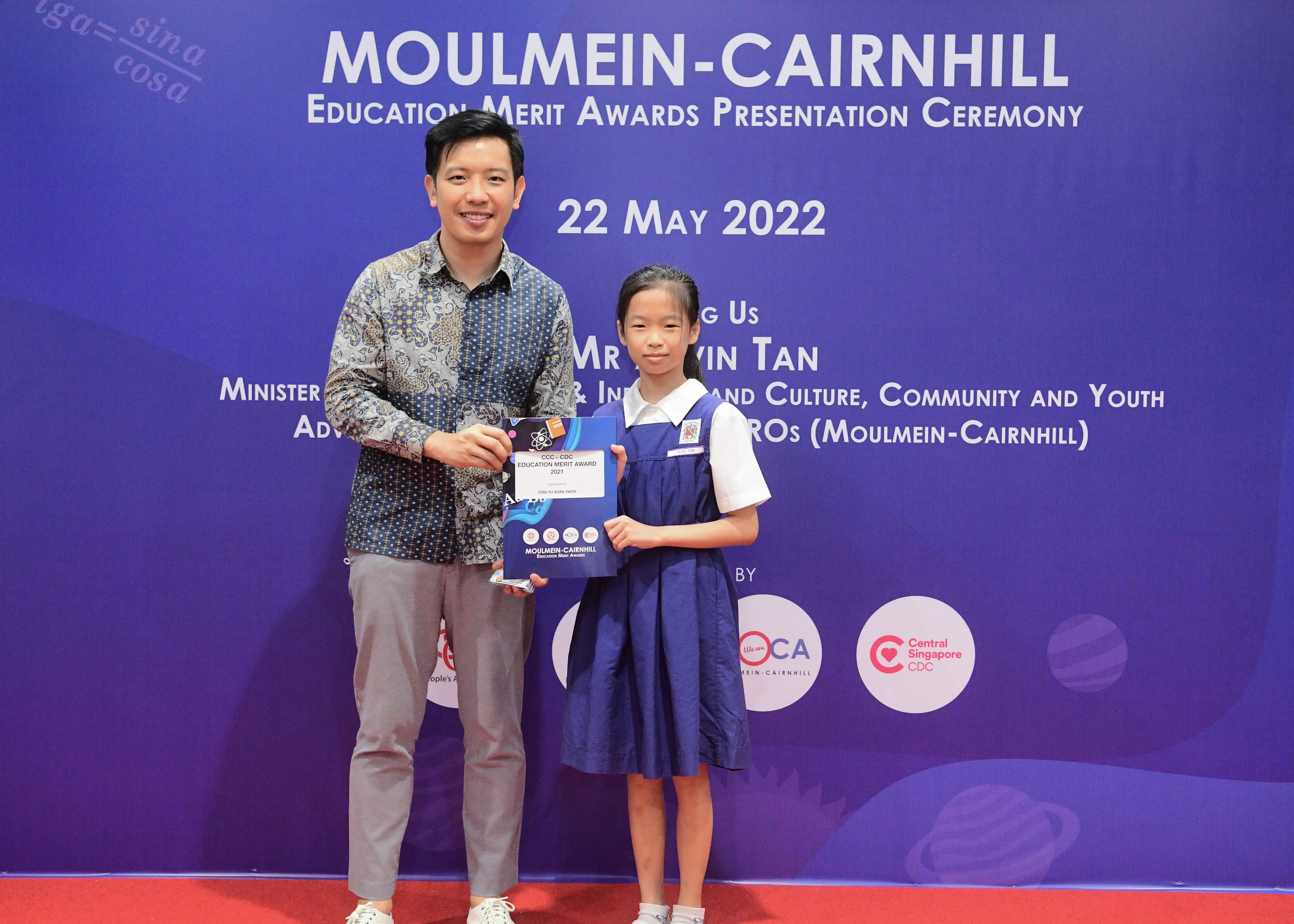 A man and a young girl in school uniform hold an award certificate at the Moulmein-Cairnhill Education Merit Awards ceremony.