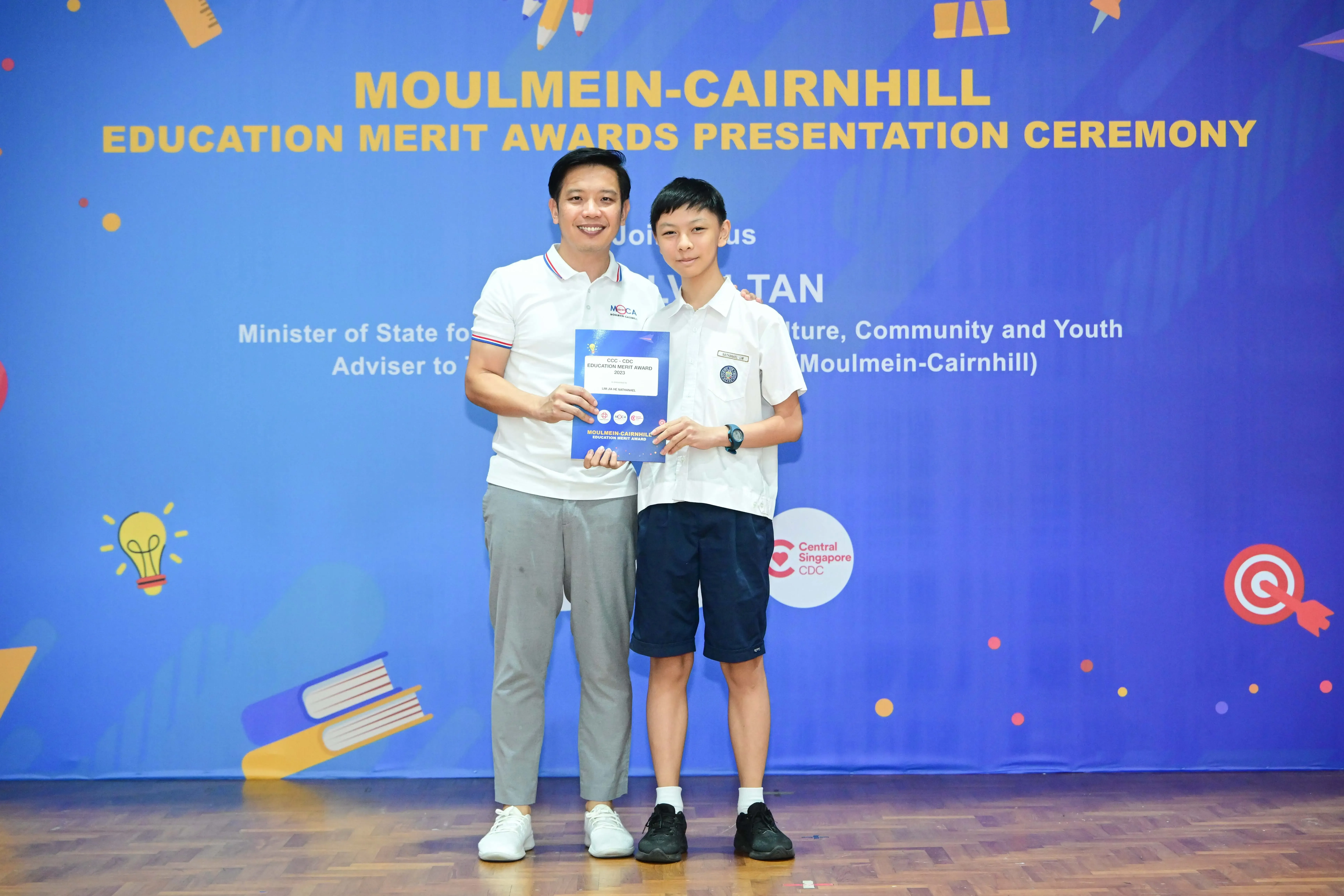 An adult and a young student hold a certificate together at the Moulmein-Cairnhill Education Merit Awards Ceremony.