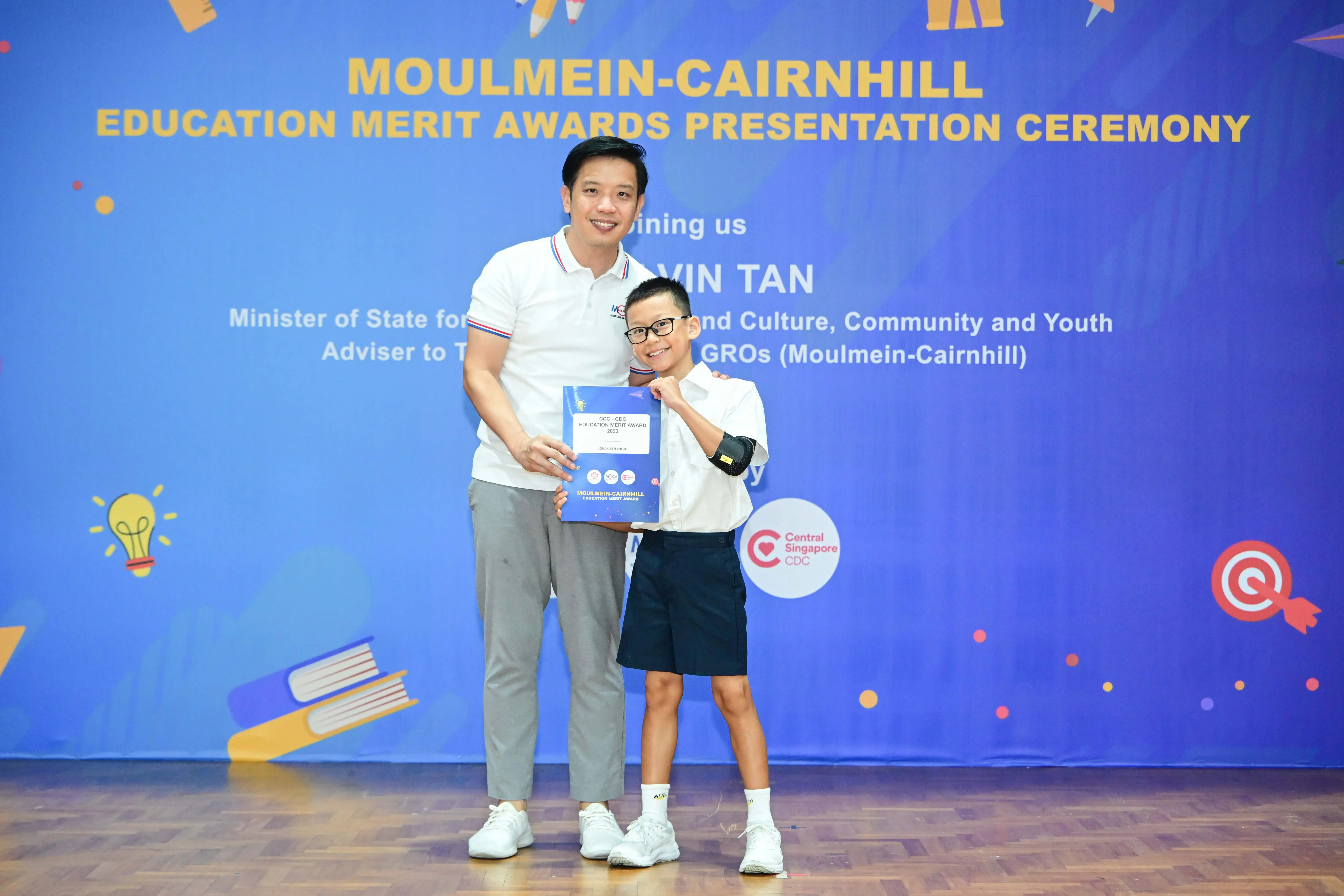 Adult and young boy holding an award certificate at Moulmein-Cairnhill Education Merit Awards Presentation Ceremony.