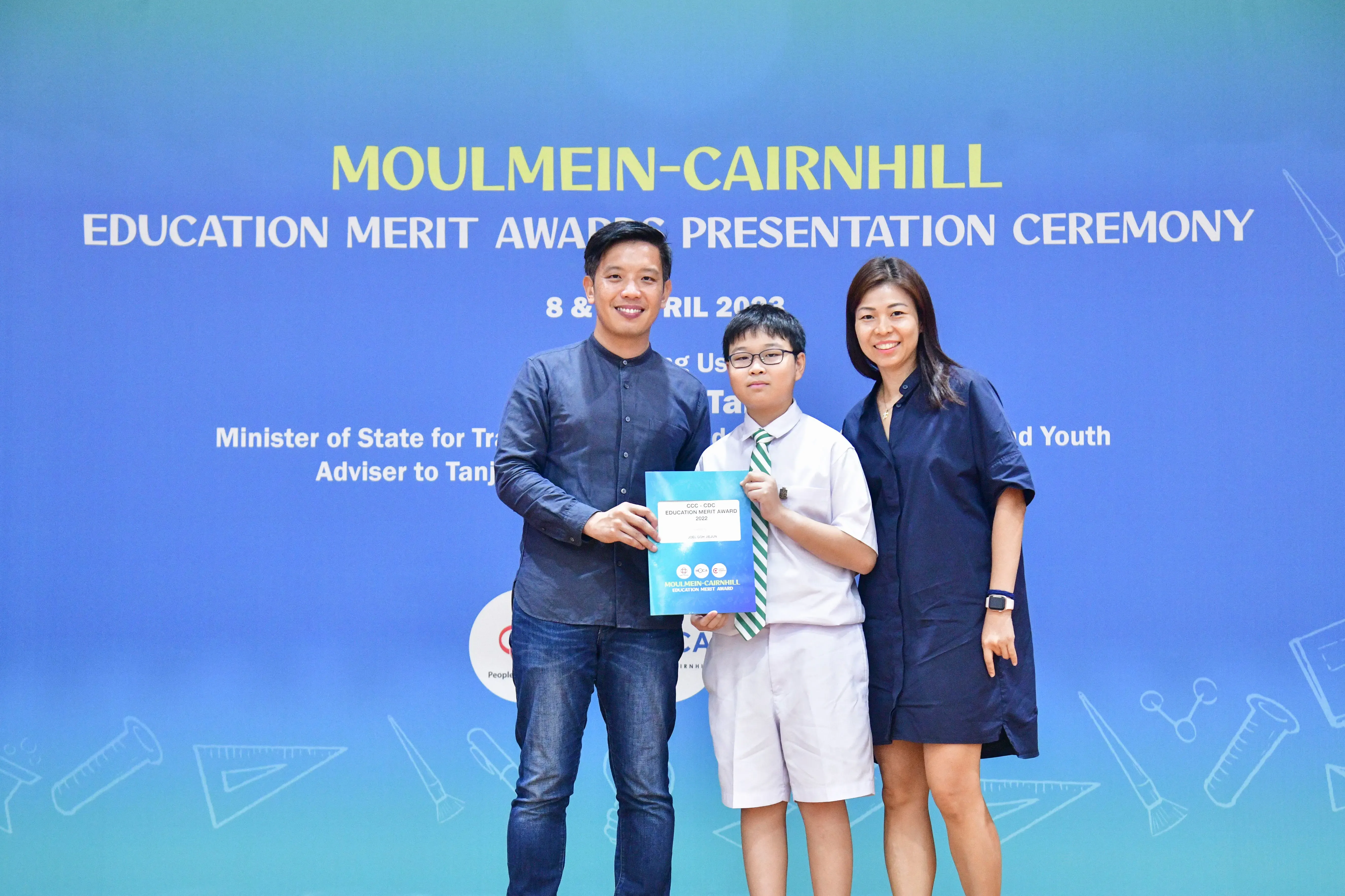 A boy in school uniform holds an award certificate flanked by two adults at the Moulmein-Cairnhill Education Merit Awards ceremony.