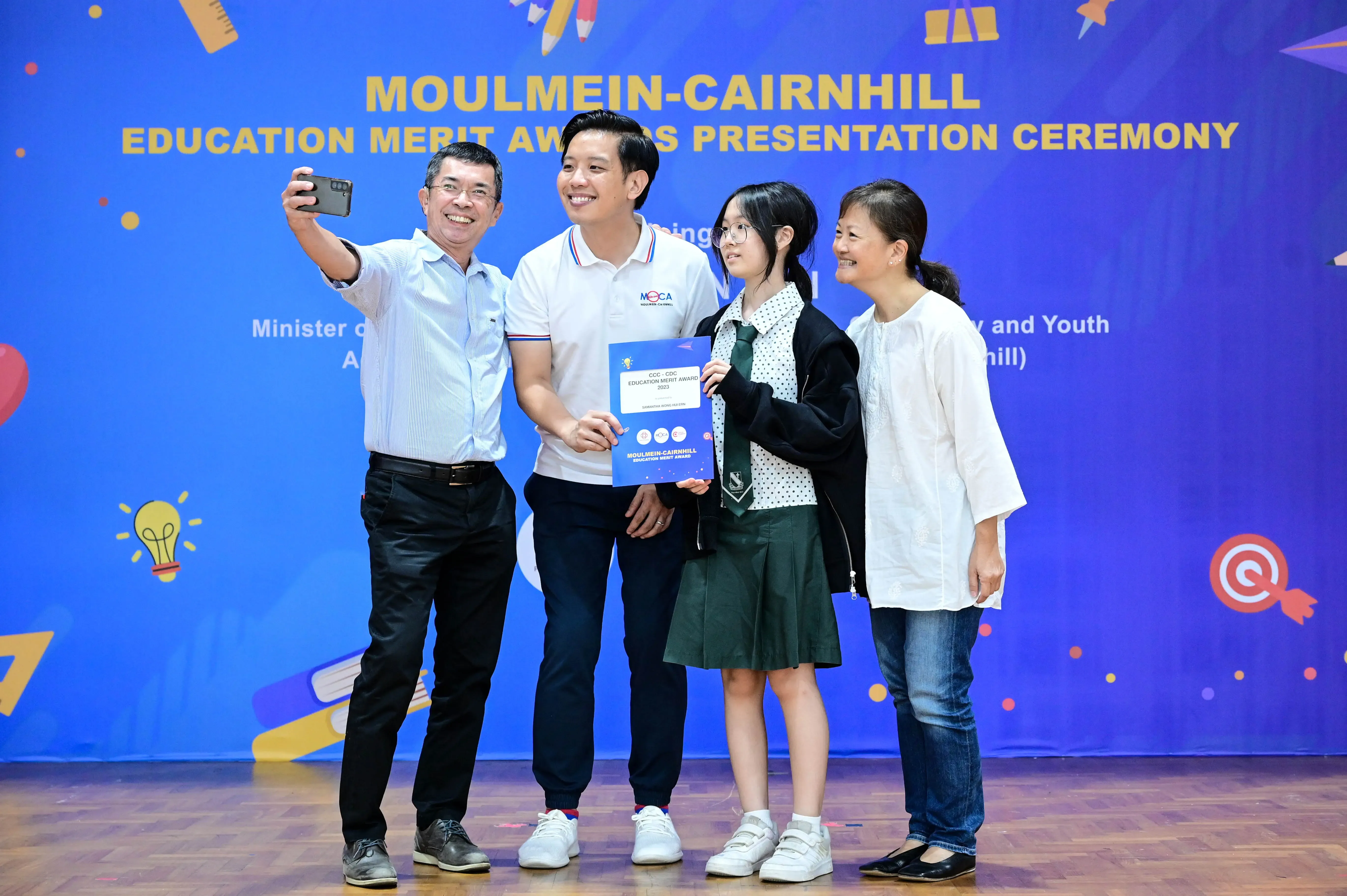 Four people take a selfie at the Moulmein-Cairnhill Education Merit Awards Presentation Ceremony, one holding a certificate.