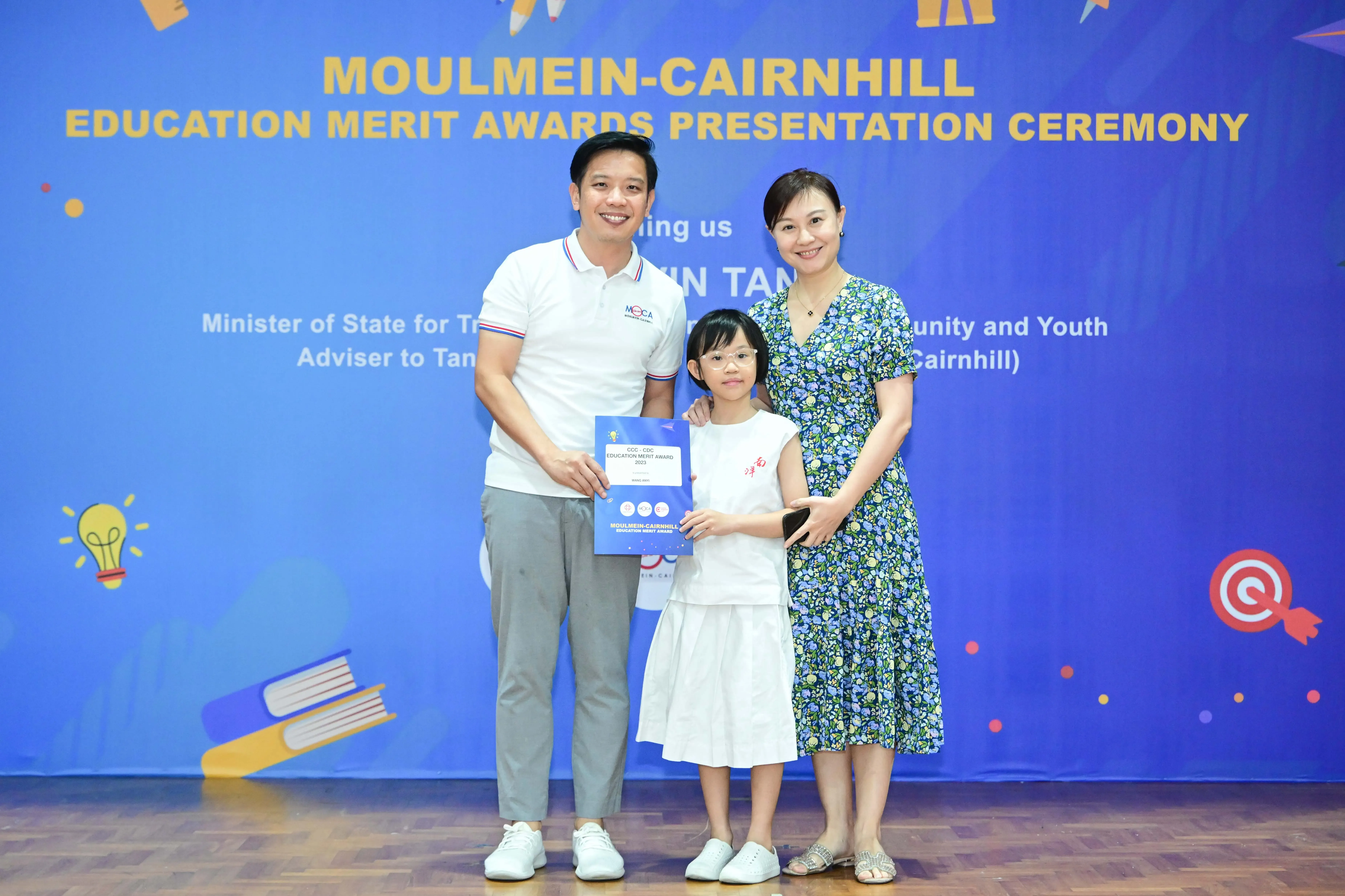 A man, young girl holding an award booklet, and woman pose before a Moulmein-Cairnhill Education Merit Awards banner.