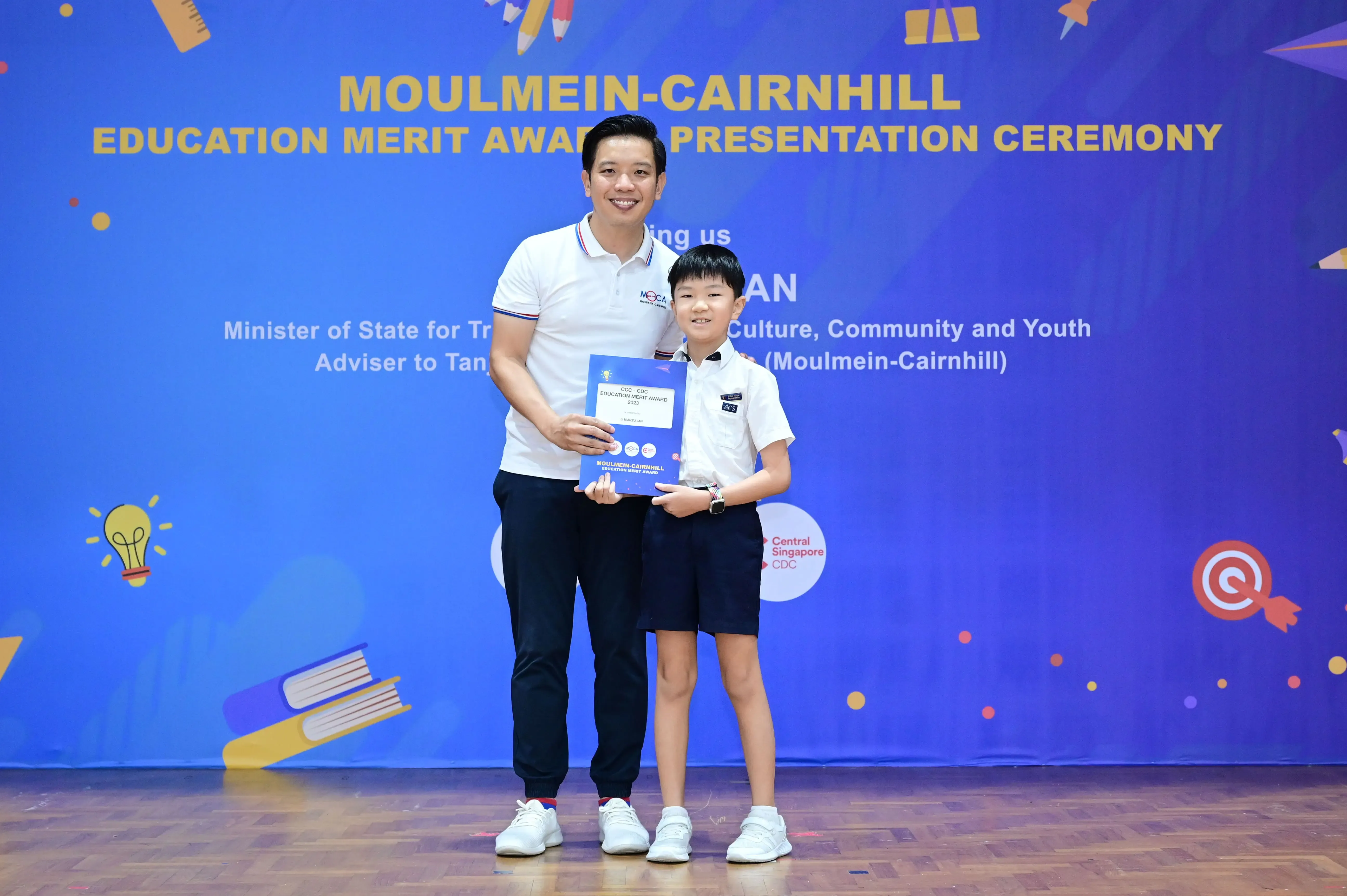 A man and a young boy hold a blue award certificate in front of a Moulmein-Cairnhill Education Merit Award banner.