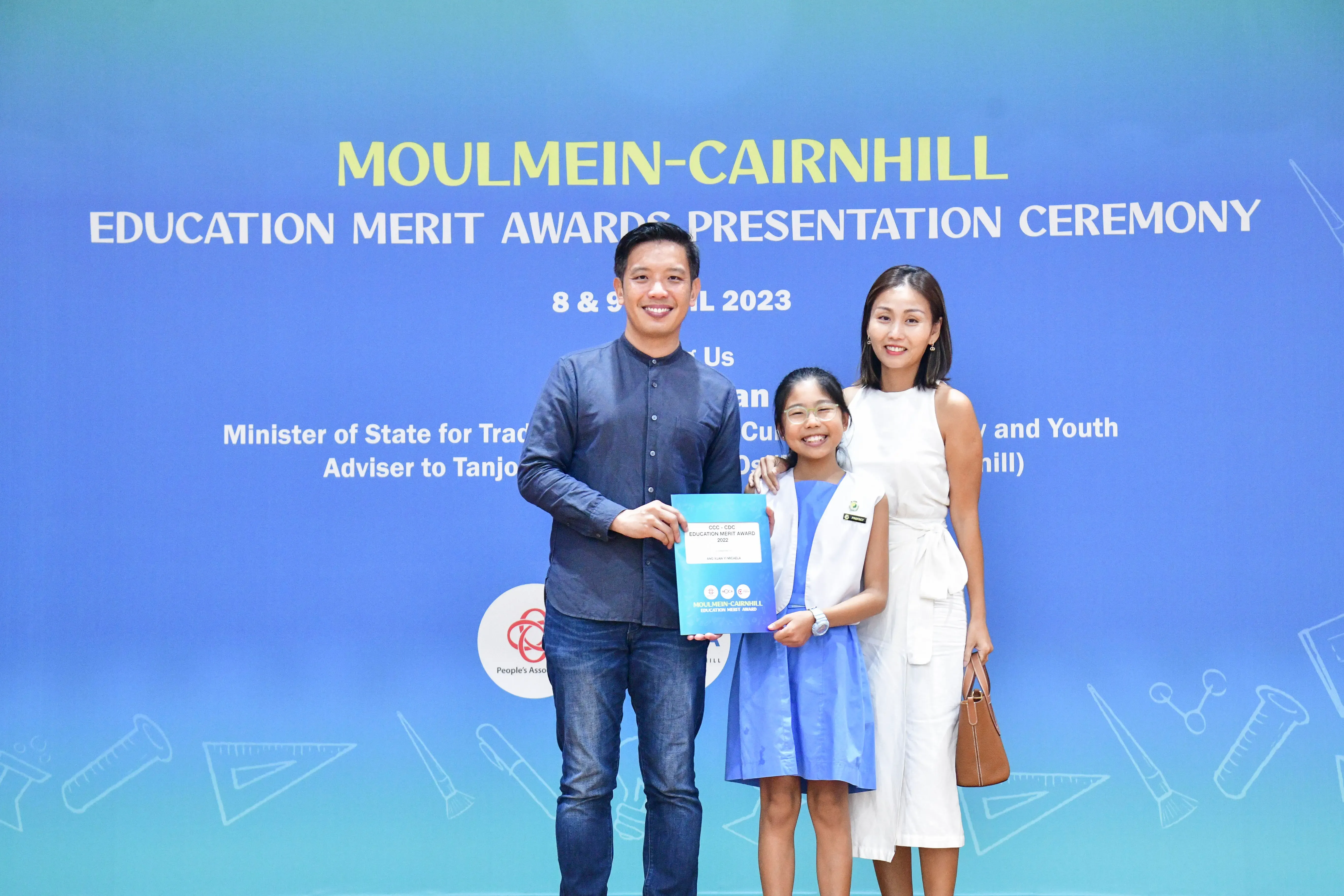 A man, young girl holding an award, and woman pose at the Moulmein-Cairnhill Education Merit Awards Ceremony 2023.