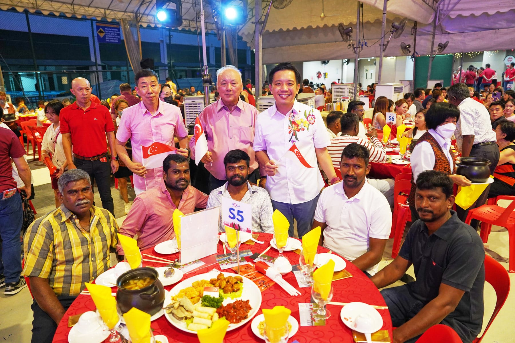 Group of people at a large outdoor banquet with red tables, food, and small Singapore flags displayed.