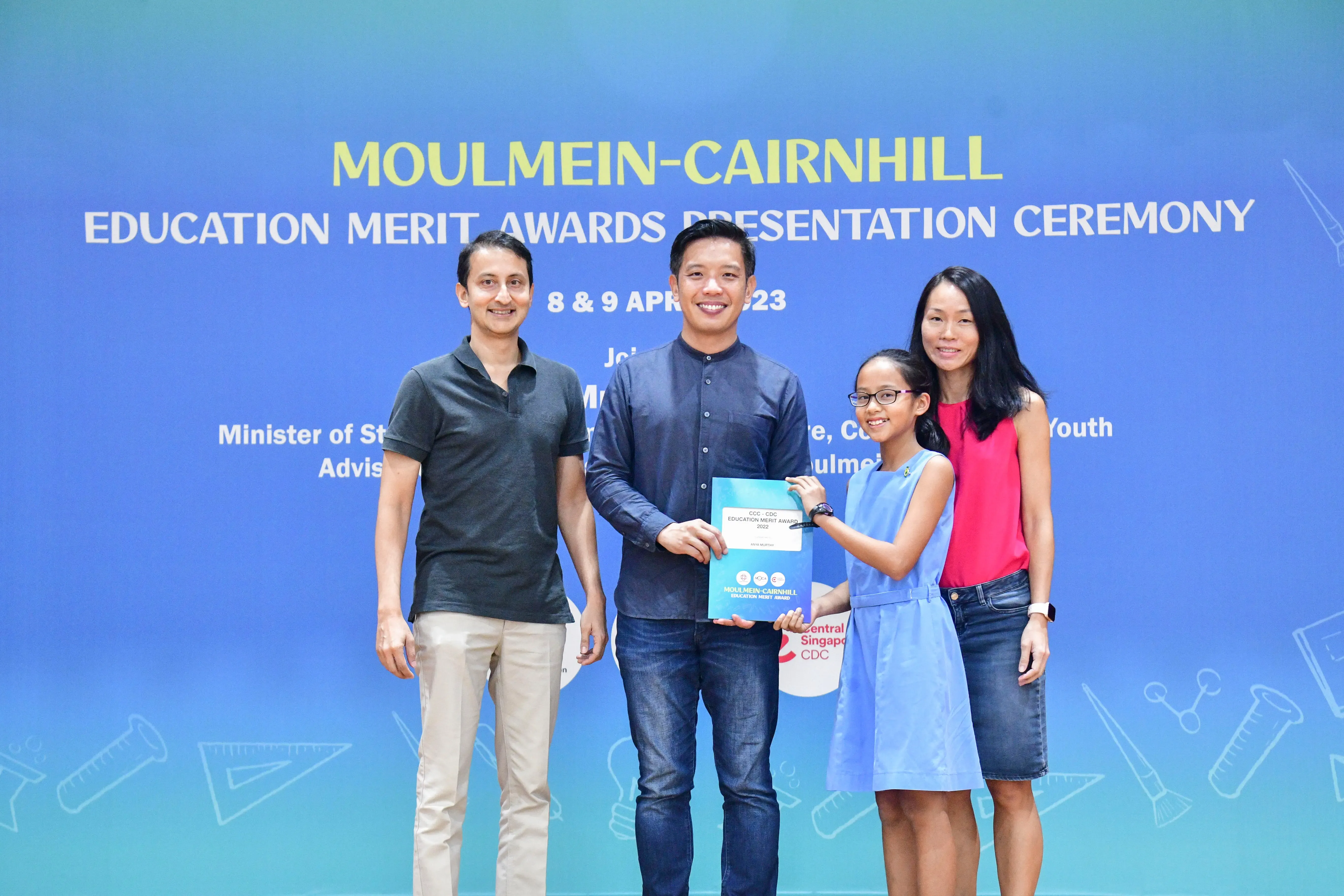 Four people pose at the Moulmein-Cairnhill Education Merit Awards Presentation Ceremony, with a girl holding an award folder.
