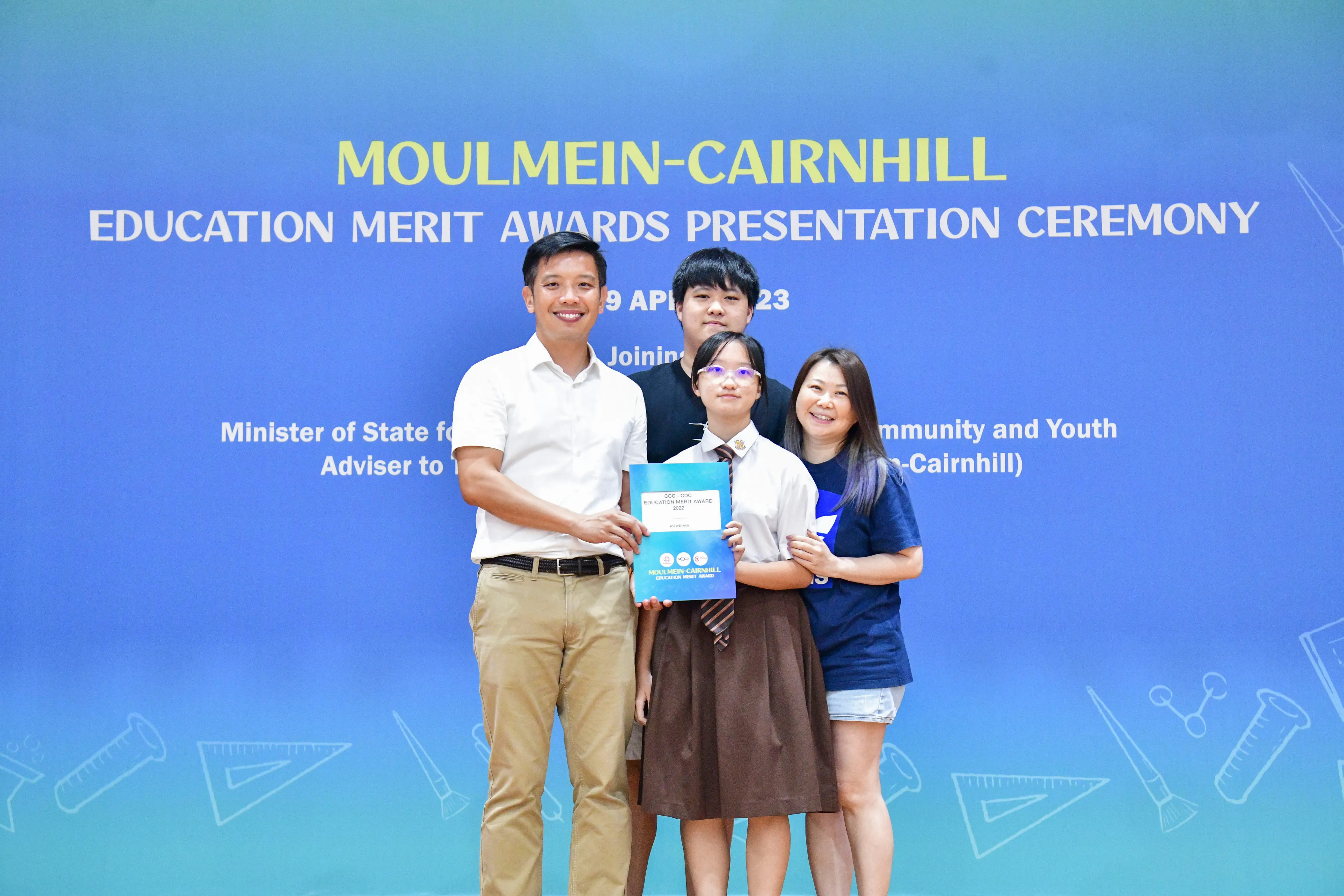 Four people pose at the Moulmein-Cairnhill Education Merit Awards Presentation Ceremony, with a student holding a blue award folder.