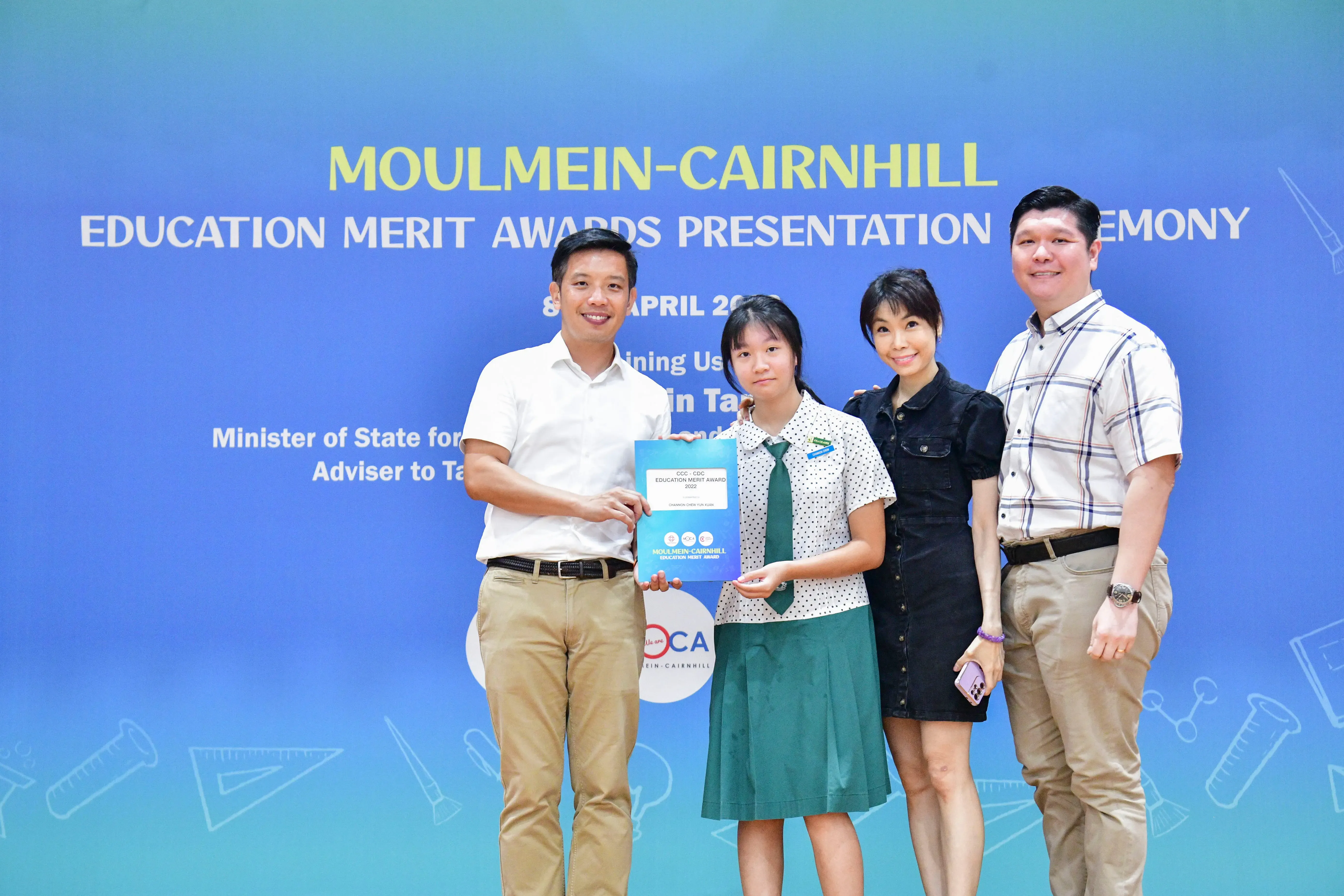 Four people pose at the Moulmein-Cairnhill Education Merit Awards Presentation Ceremony, with a student holding an award folder.