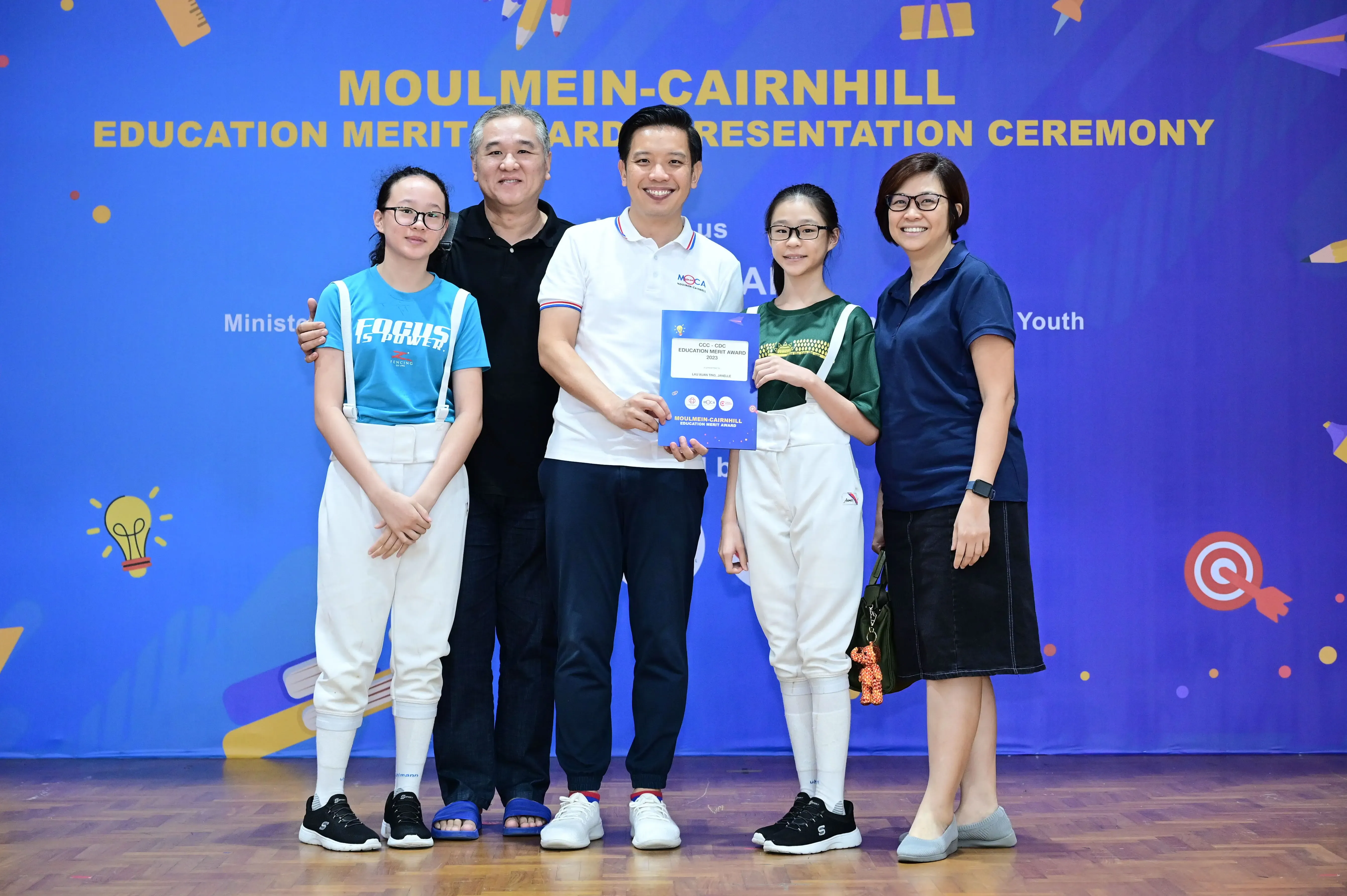 Five people pose with a certificate at the Moulmein-Cairnhill Education Merit Award Presentation Ceremony banner.