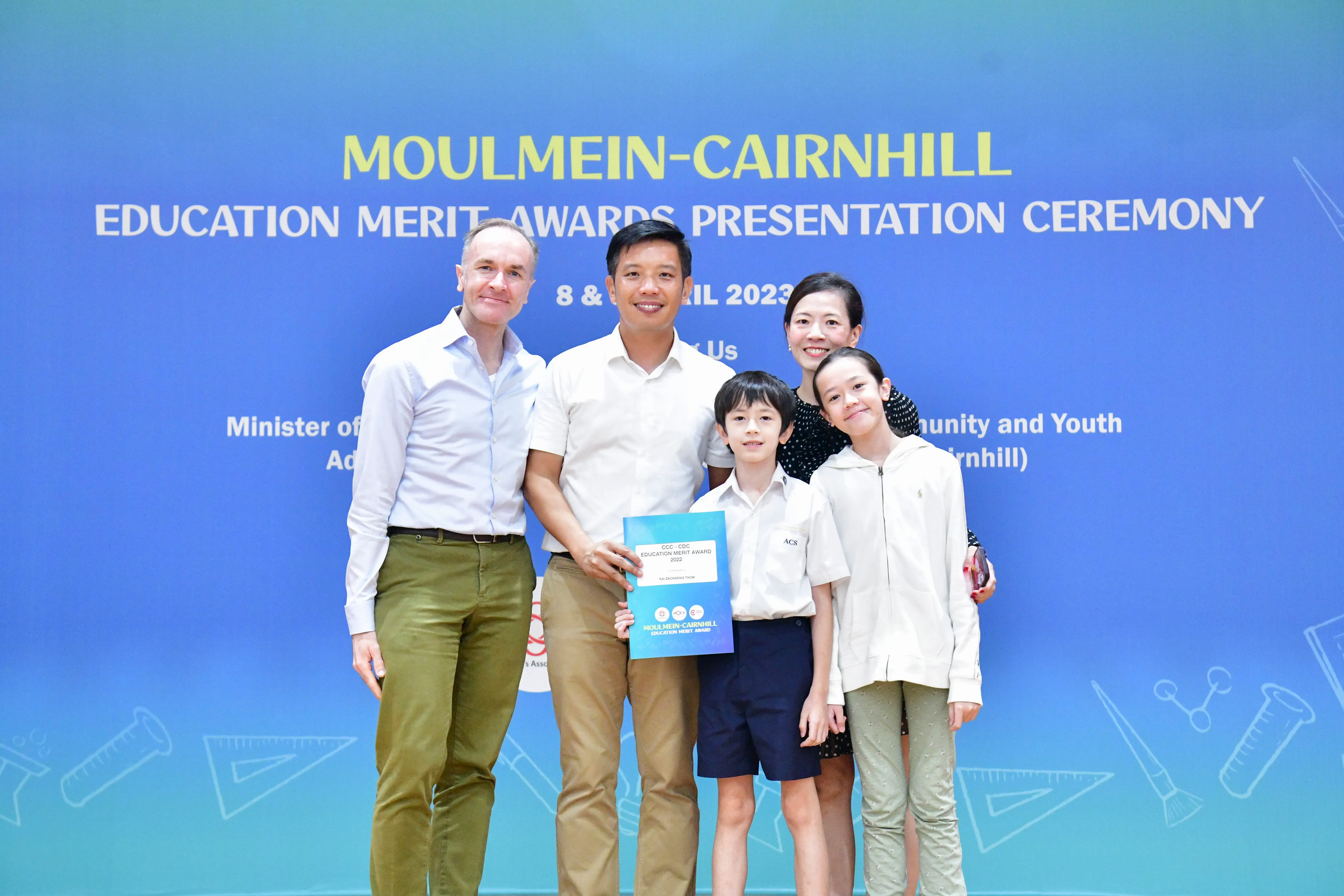 Five people pose in front of a blue Moulmein-Cairnhill Education Merit Awards Presentation Ceremony backdrop, holding an award folder.