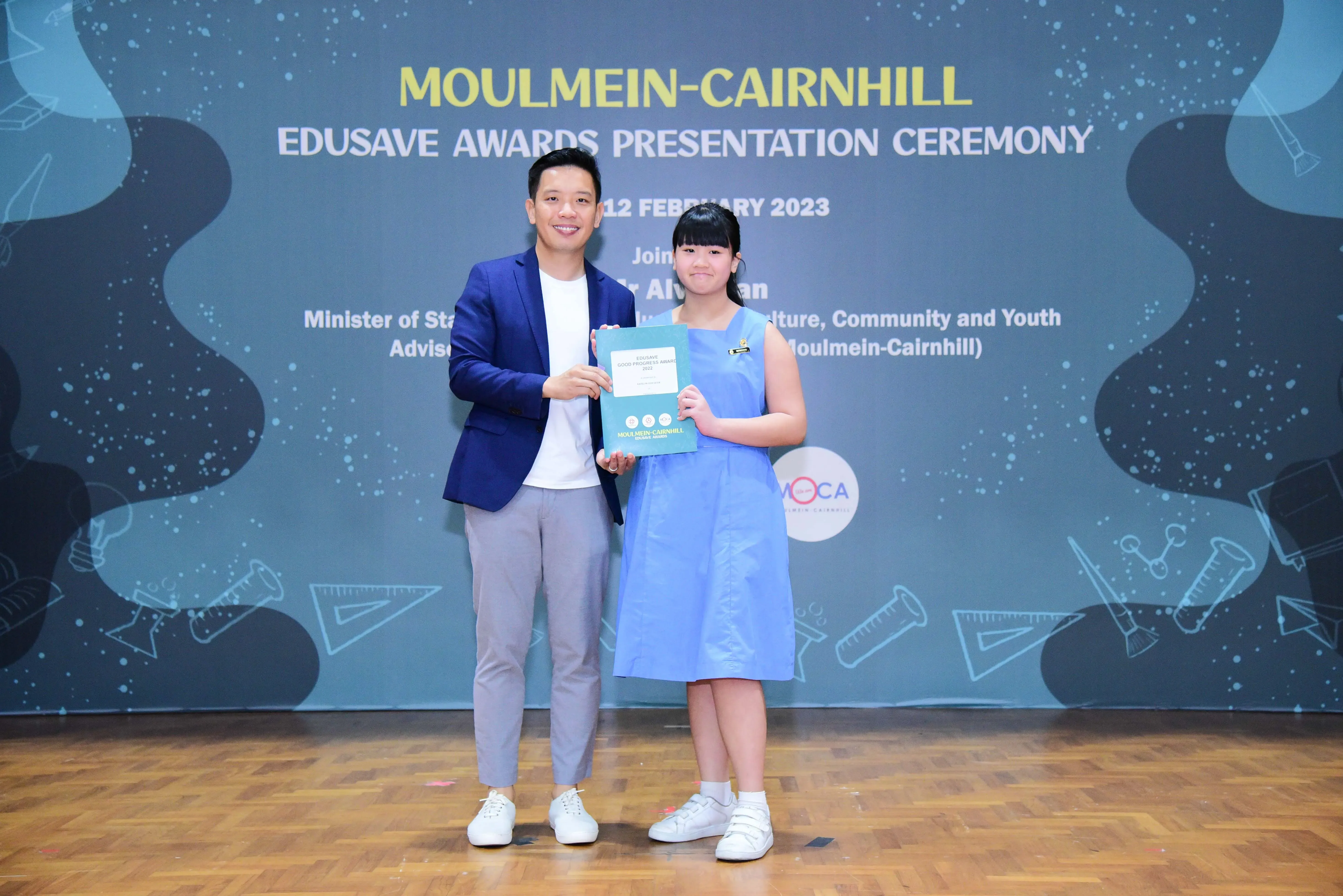 A man in a blue blazer and a girl in a blue dress hold a certificate at the Moulmein-Cairnhill Edusave Awards Ceremony.