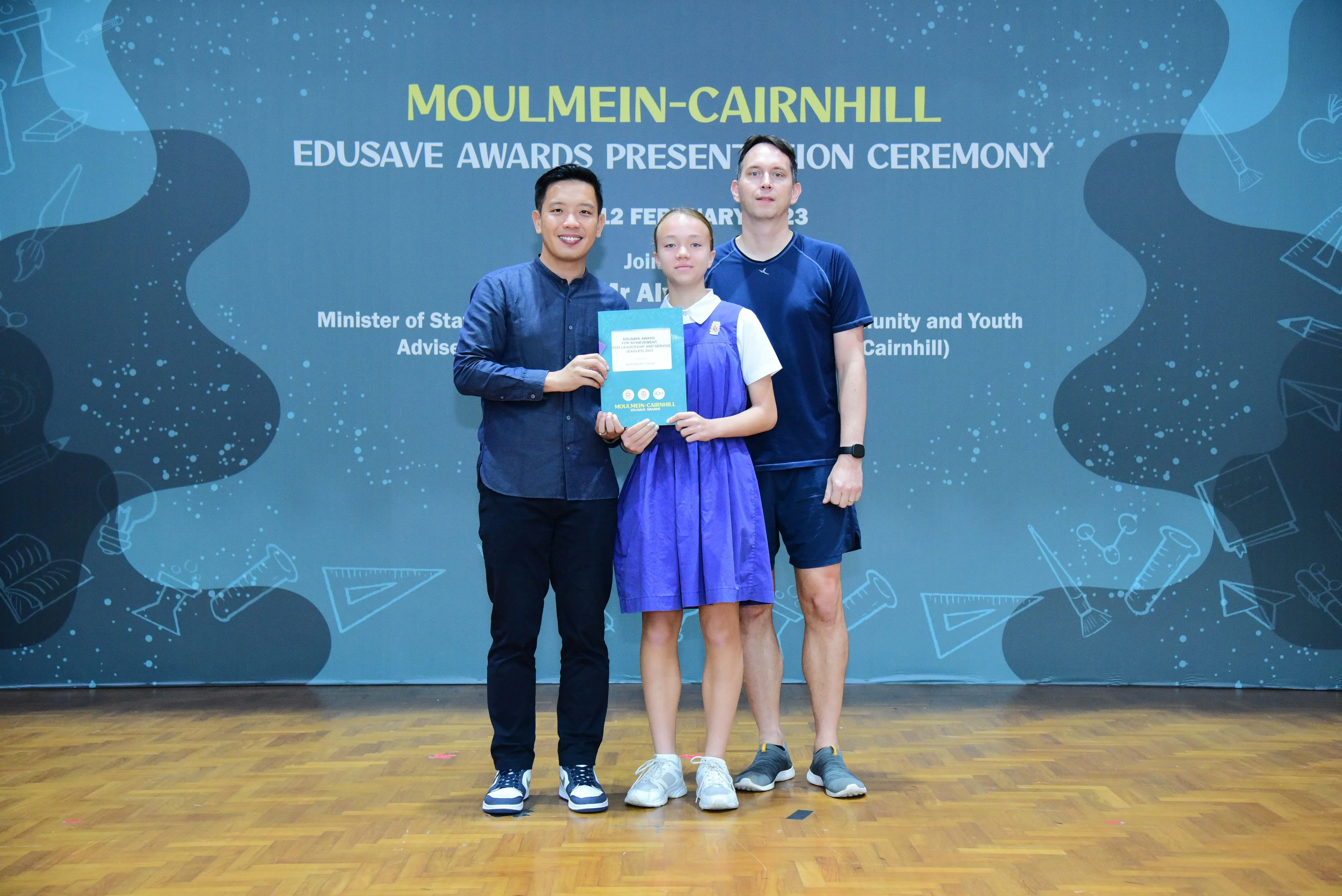 Three people pose at the Moulmein-Cairnhill Edusave Awards Ceremony, with a girl in blue holding a certificate.