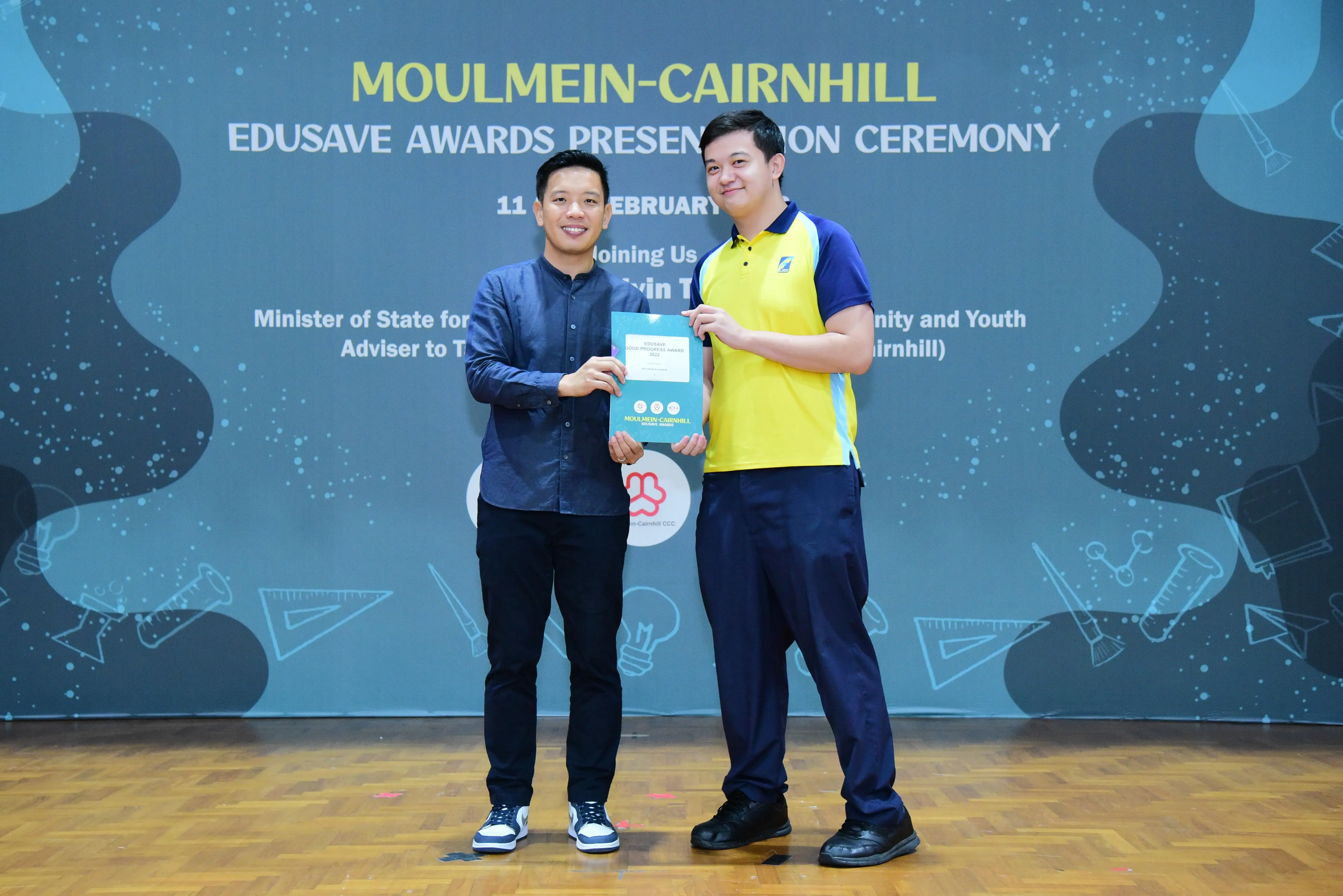 Two men hold a certificate together at the Moulmein-Cairnhill Edusave Awards Presentation Ceremony backdrop.