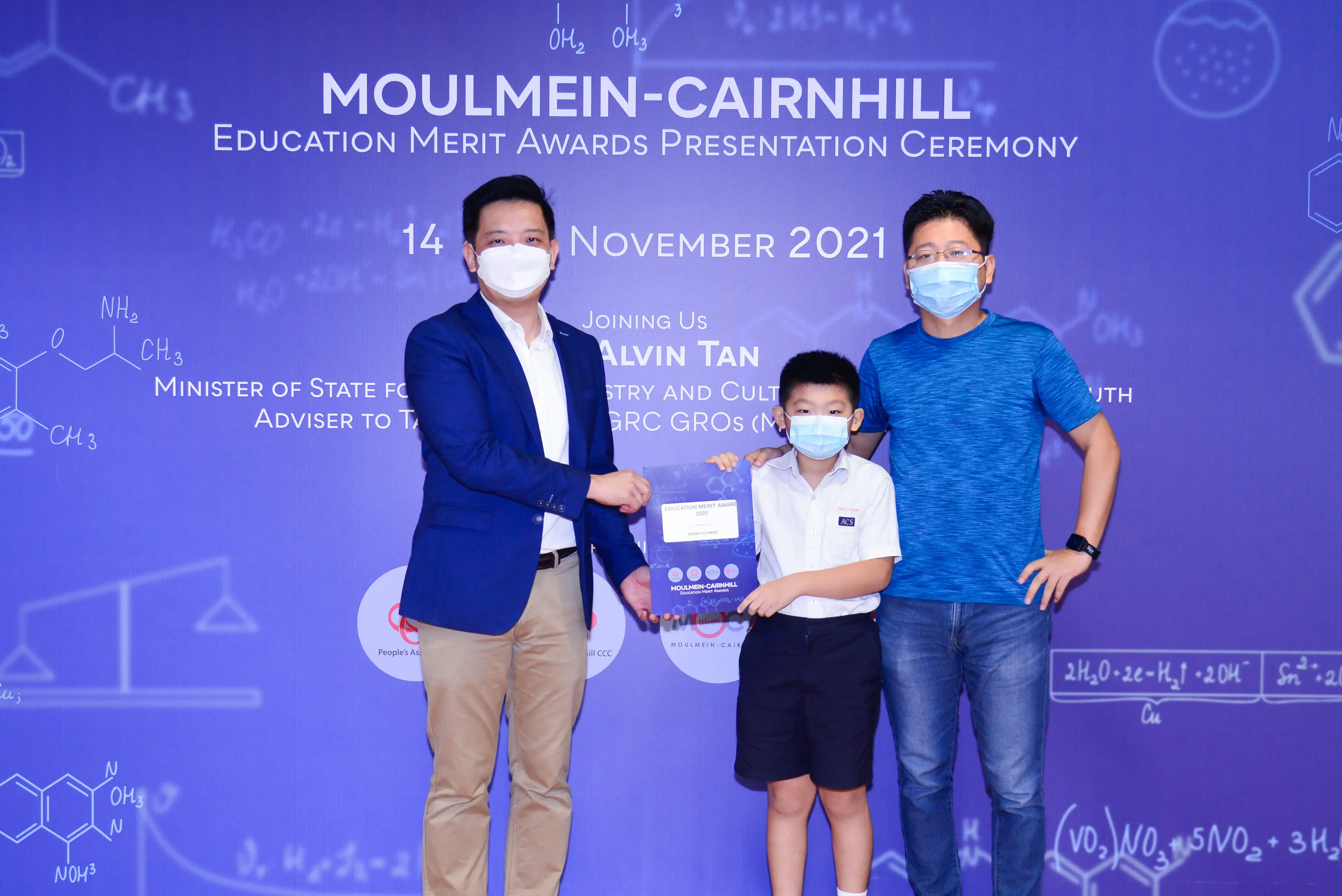 Two masked men and a young boy hold an award certificate at the Moulmein-Cairnhill Education Merit Awards ceremony.
