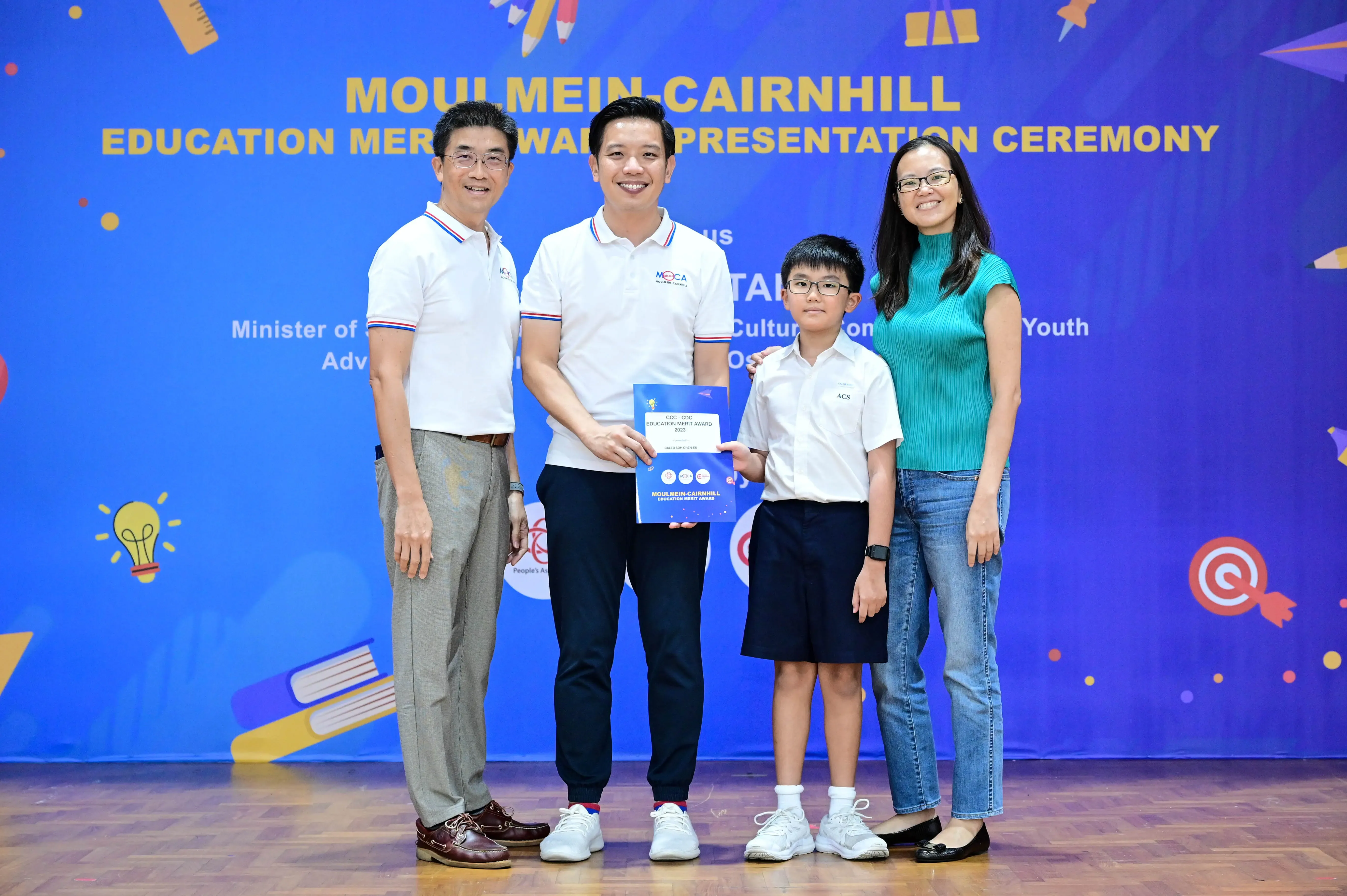 Four people pose at the Moulmein-Cairnhill Education Merit Award Presentation Ceremony, holding a blue award booklet.