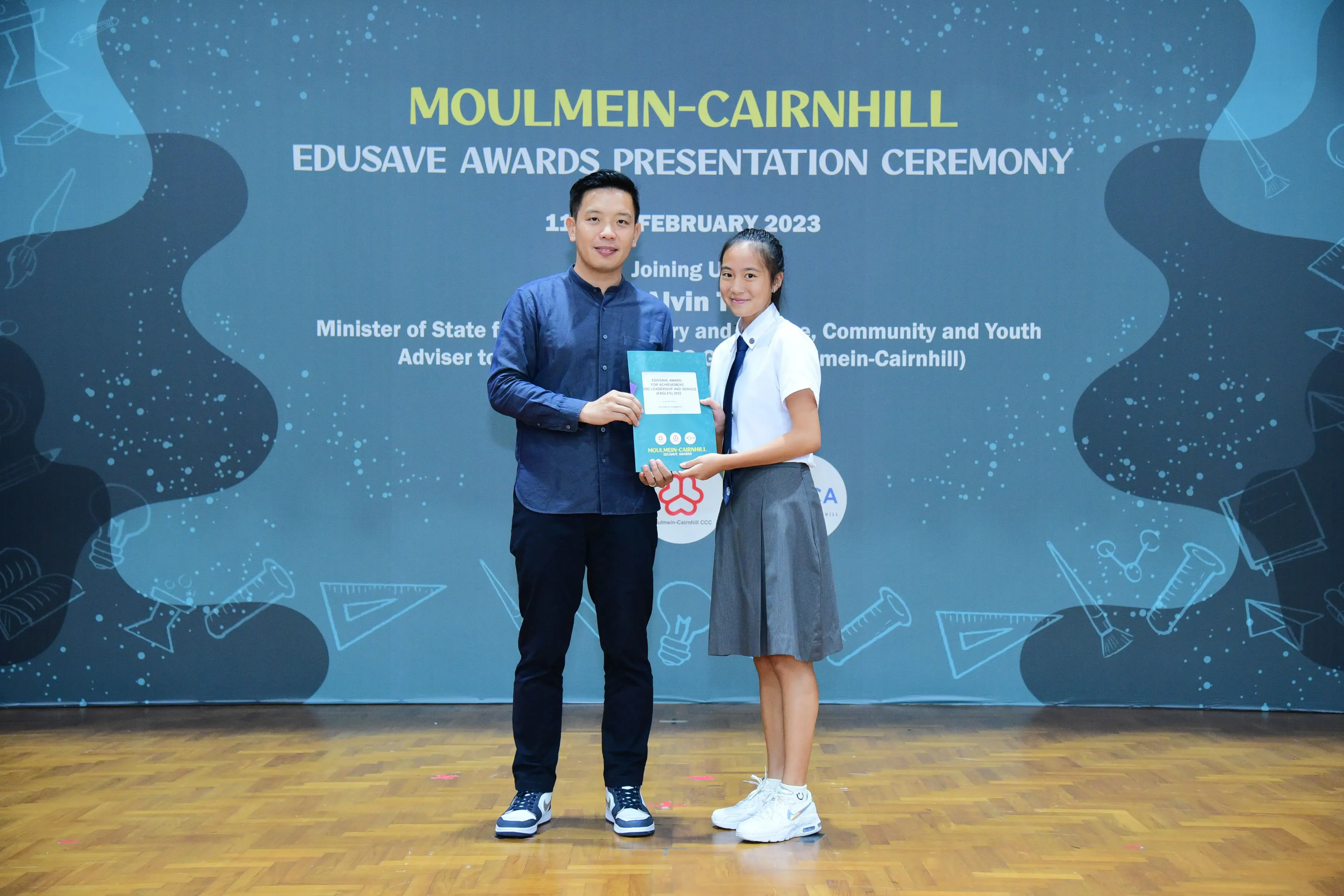 A man and a young girl in school uniform hold an award together at the Moulmein-Cairnhill Edusave Awards Ceremony 2023.