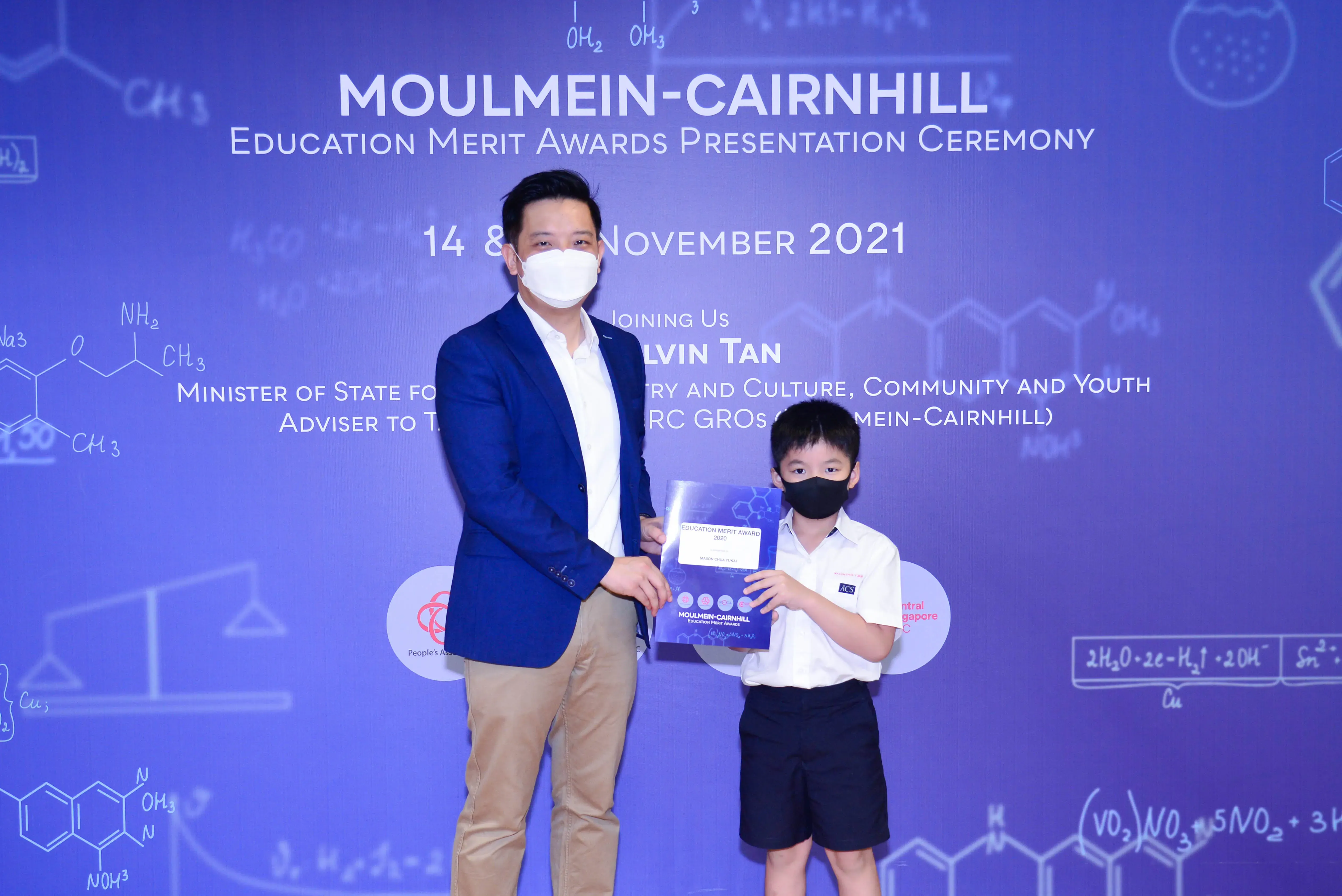 An adult in a blue blazer and a young boy in school uniform hold an award at the Moulmein-Cairnhill Education Merit Awards 2021.