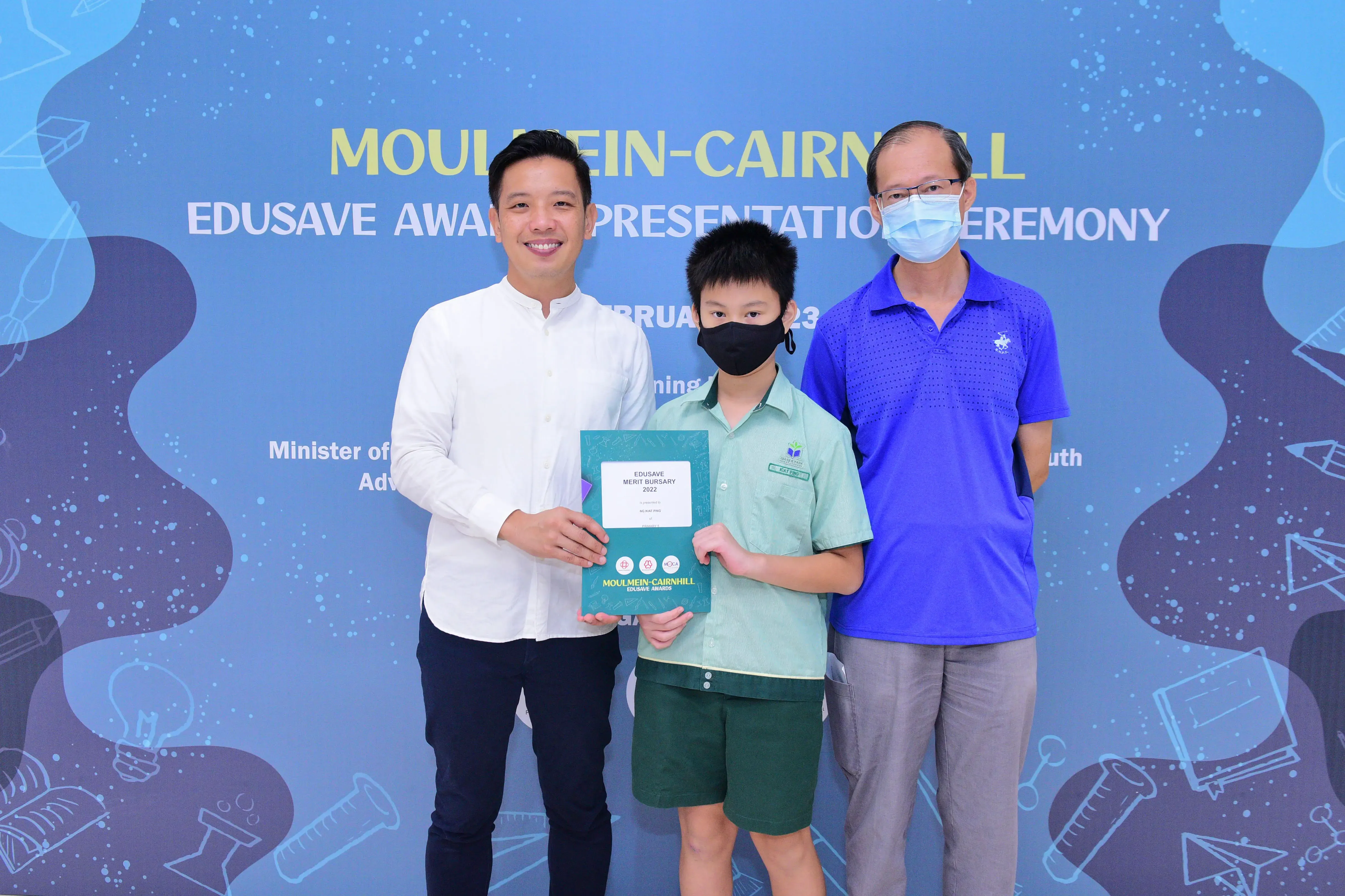 A boy holding an Edusave Merit Bursary certificate stands between two adults at the Moulmein-Cairnhill award ceremony.