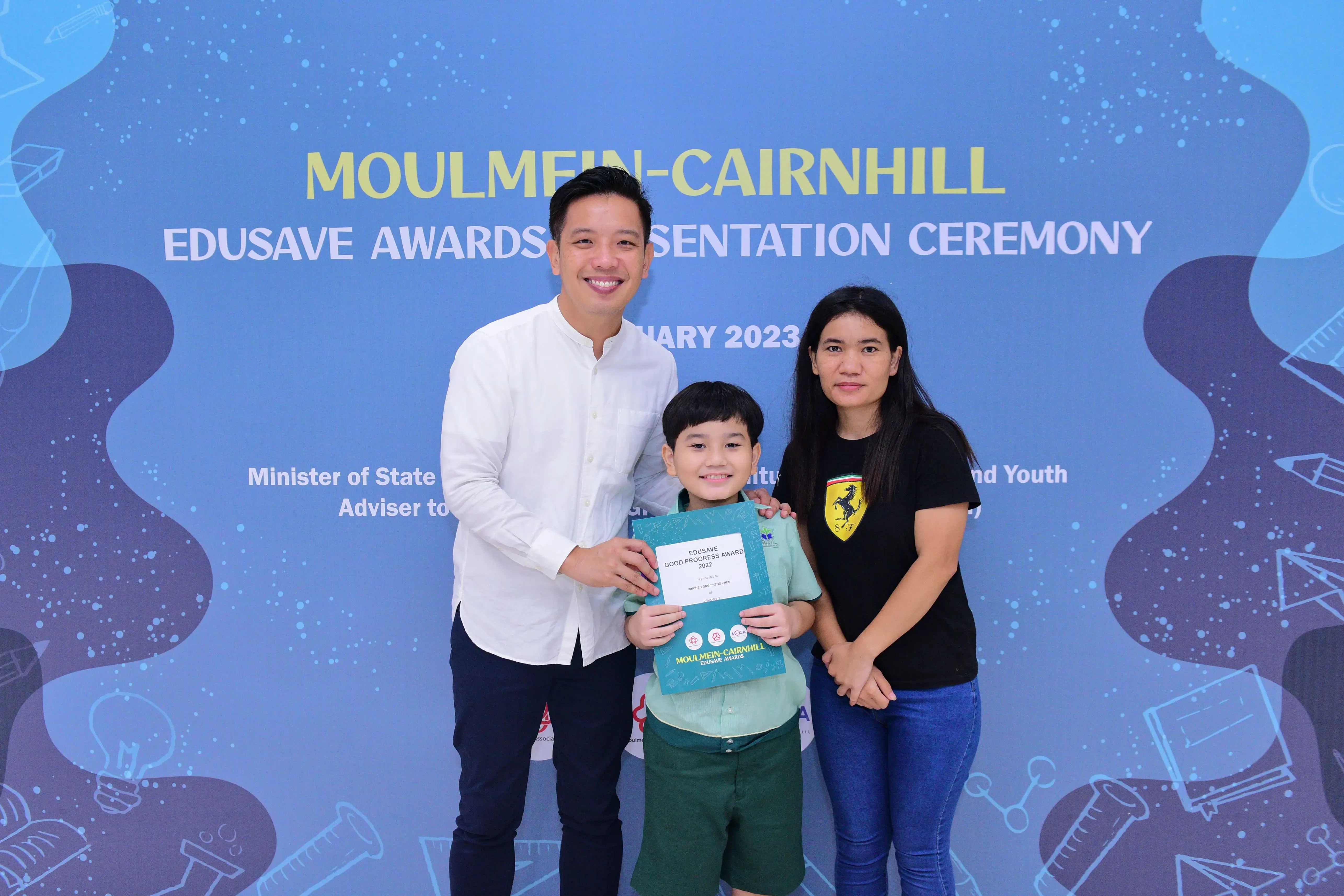 A boy holds an Edusave award certificate flanked by a man and woman at the Moulmein-Cairnhill ceremony, January 2023.